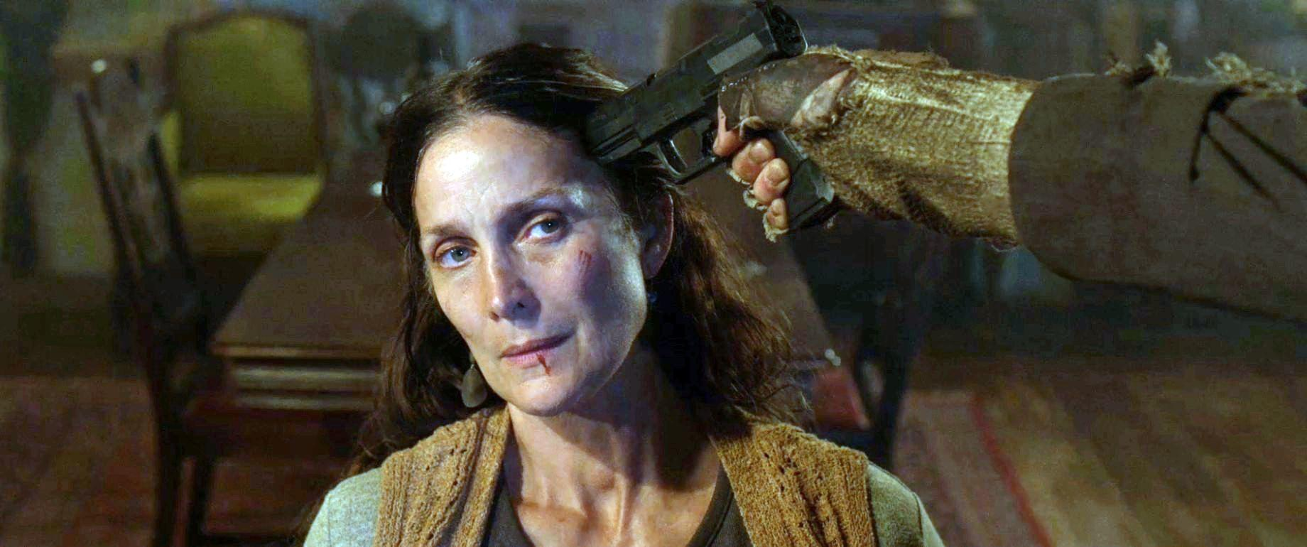 Eine Frau wird mit vorgehaltener Waffe festgehalten. Eine Hand mit einer Pistole wird gegen ihren Kopf gedrückt. Sie hat eine blutige Lippe und eine Wunde im Gesicht.