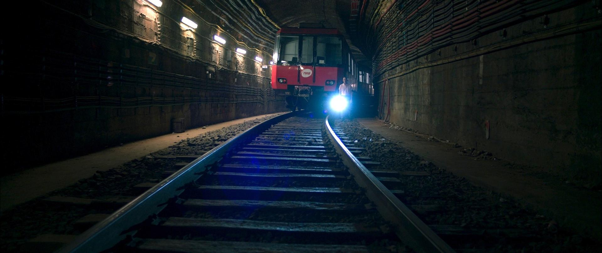 Ein roter Zug kommt aus einem dunklen Tunnel. Gleise führen zu den hellen Scheinwerfern des Zuges. Wände sind dunkel und mit Kabeln versehen.