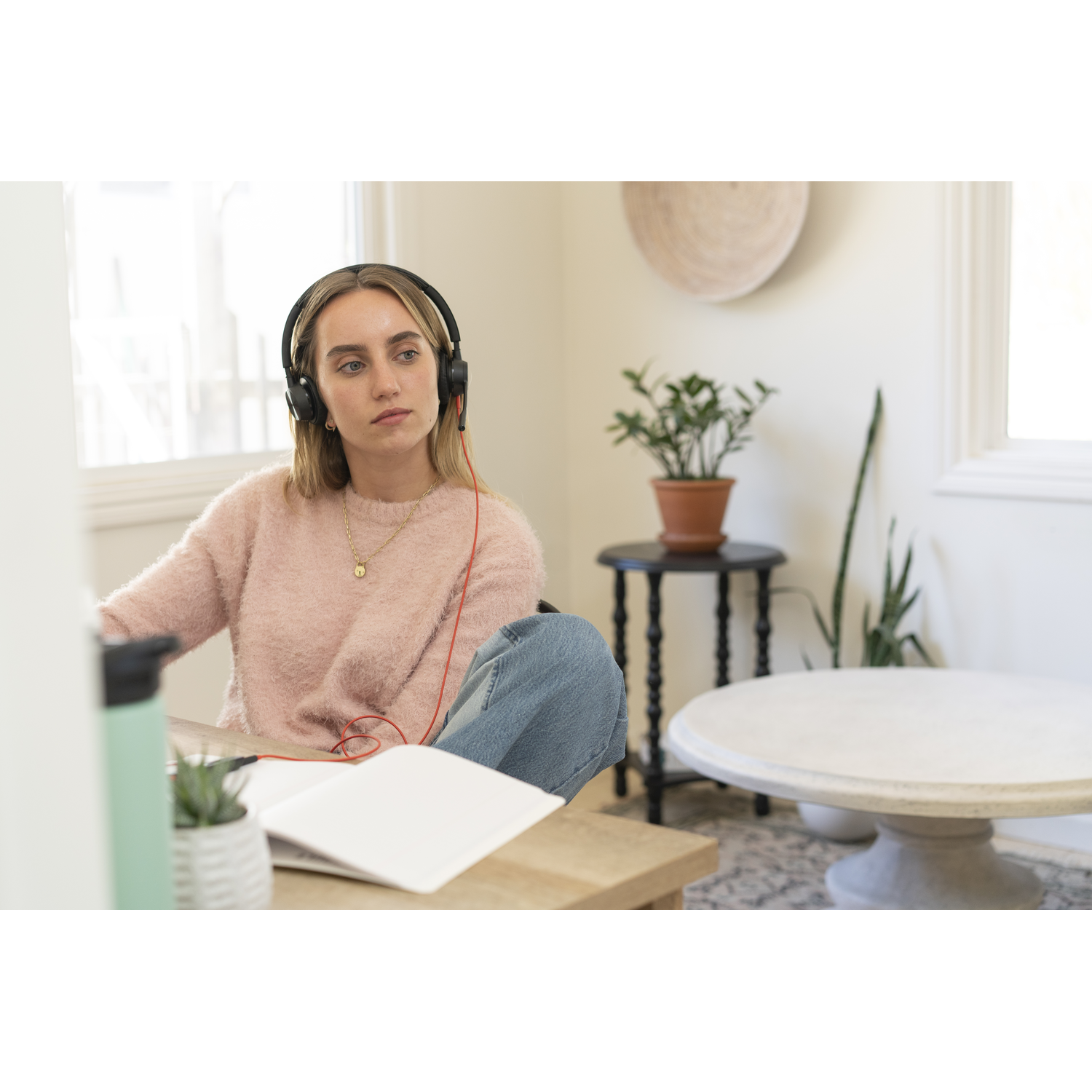 A woman with headphones sits at a table, looking away. She wears a pink sweater and jeans. A notebook and water bottle are present.