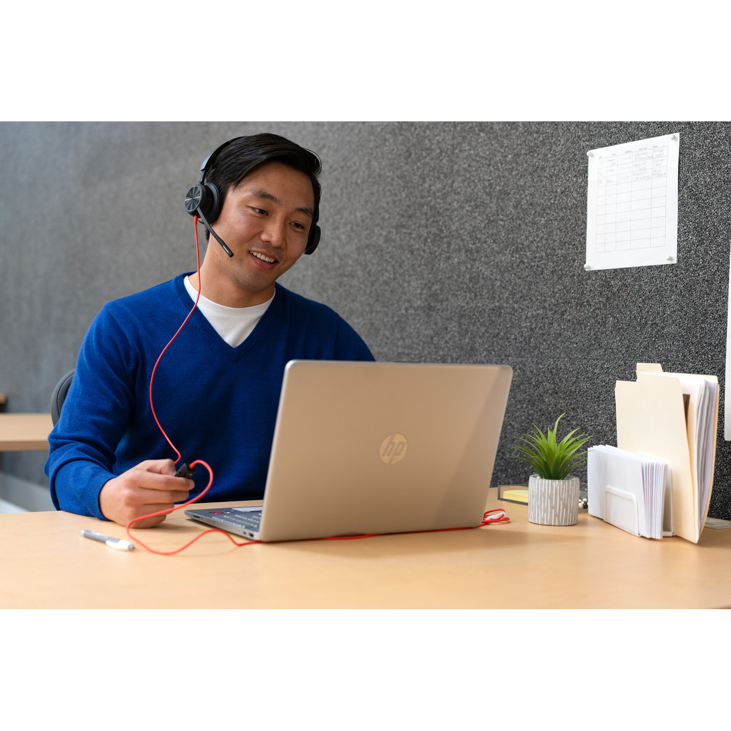 A man wearing a headset is using a laptop. He is in an office setting. A plant and files are on the desk.