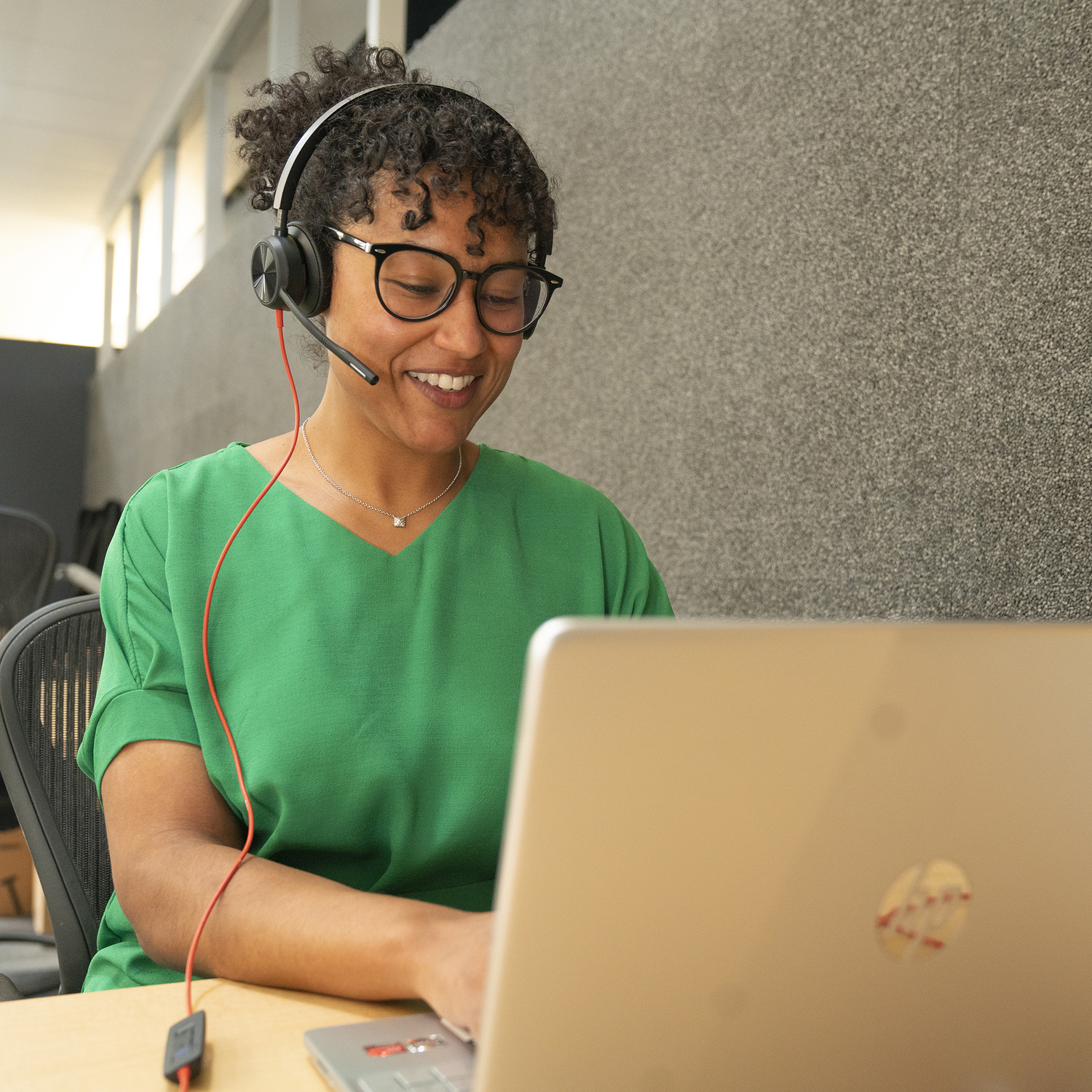 A smiling woman wearing glasses and a headset works on a laptop. She wears a green shirt, and a red cable runs down.