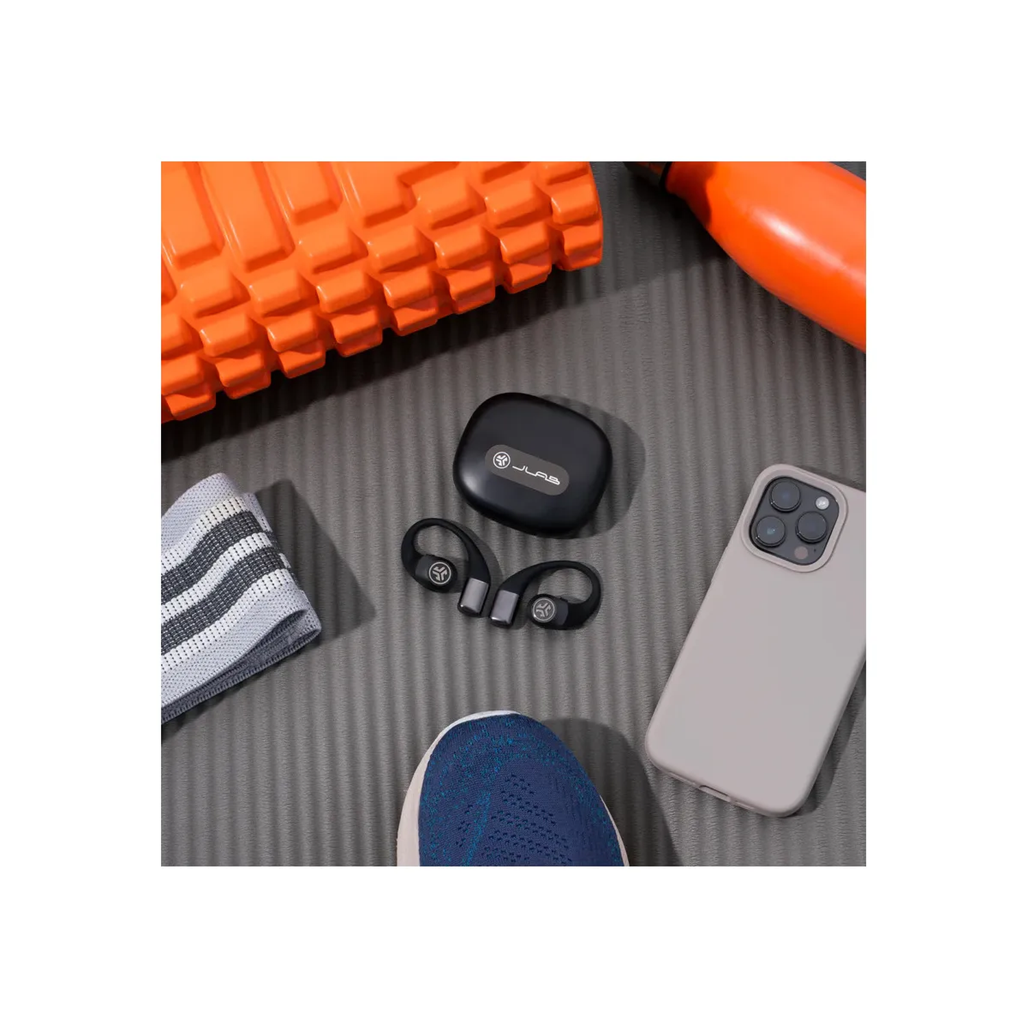 Overzicht met fitnessapparatuur: oranje foamroller, zwarte case met oortelefoons, telefoon, schoenen en weerstandsband.