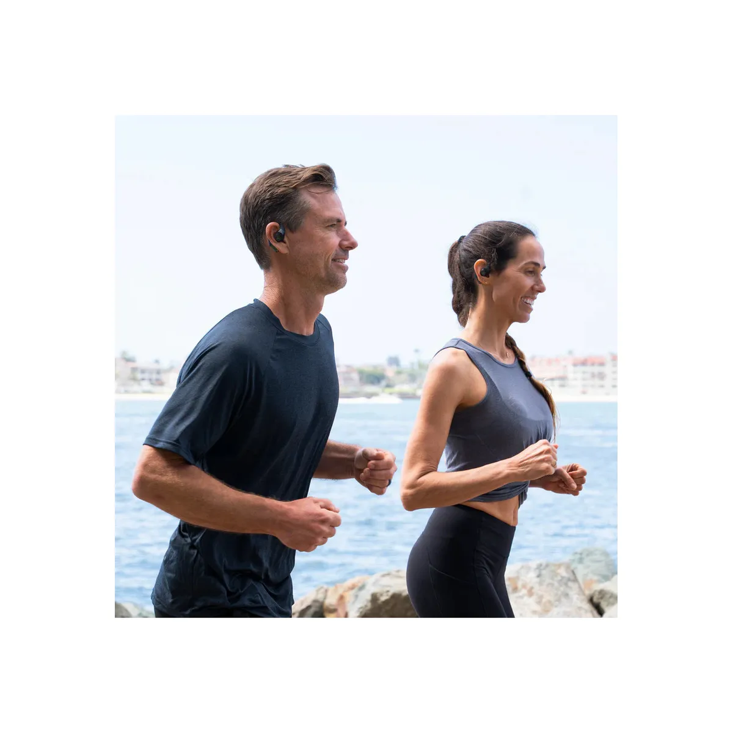 Een man en een vrouw rennen naast elkaar bij de waterkant. Ze dragen sportkleding en oortelefoons. De zee en een stad zijn op de achtergrond.
