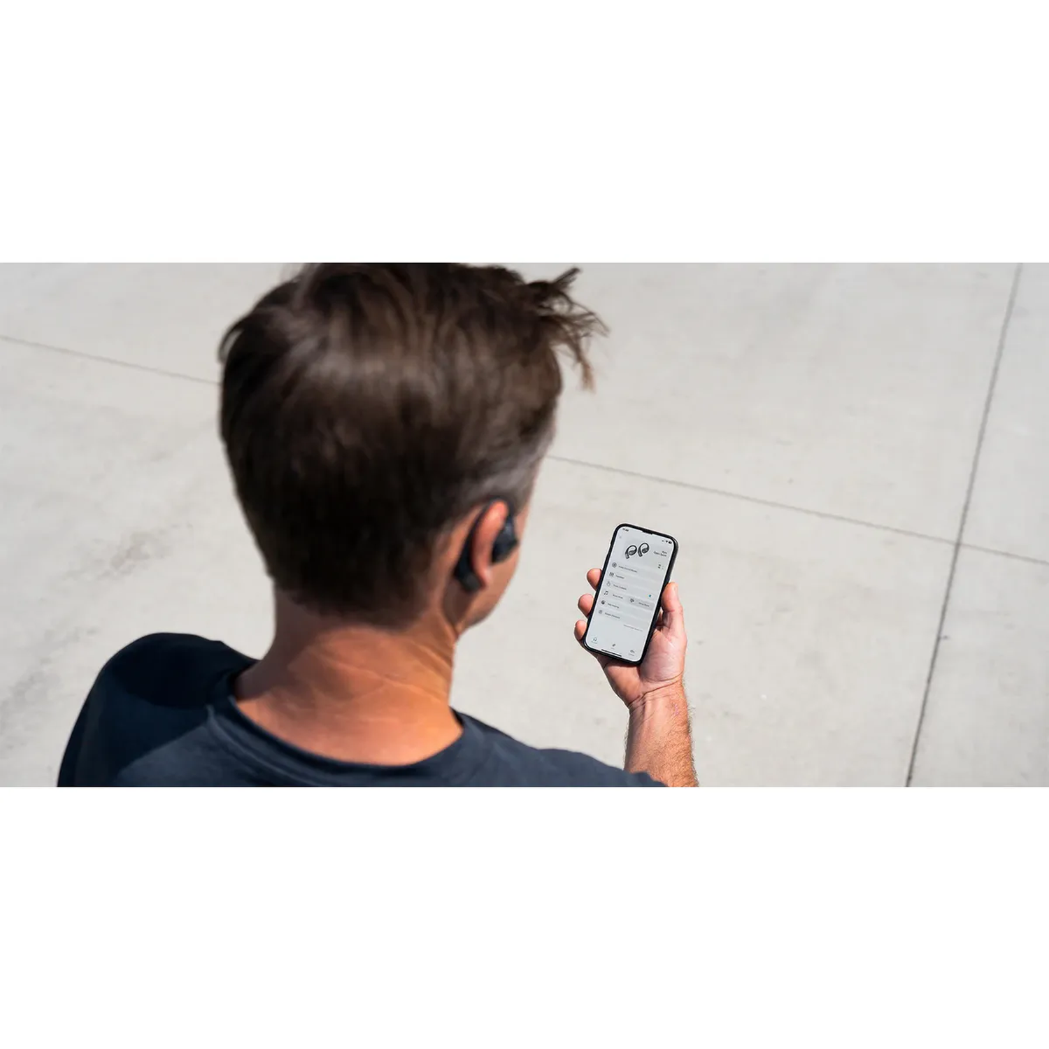 Man houdt telefoon met geopende app vast, draagt oortjes. Achtergrond is grijs betonoppervlak.