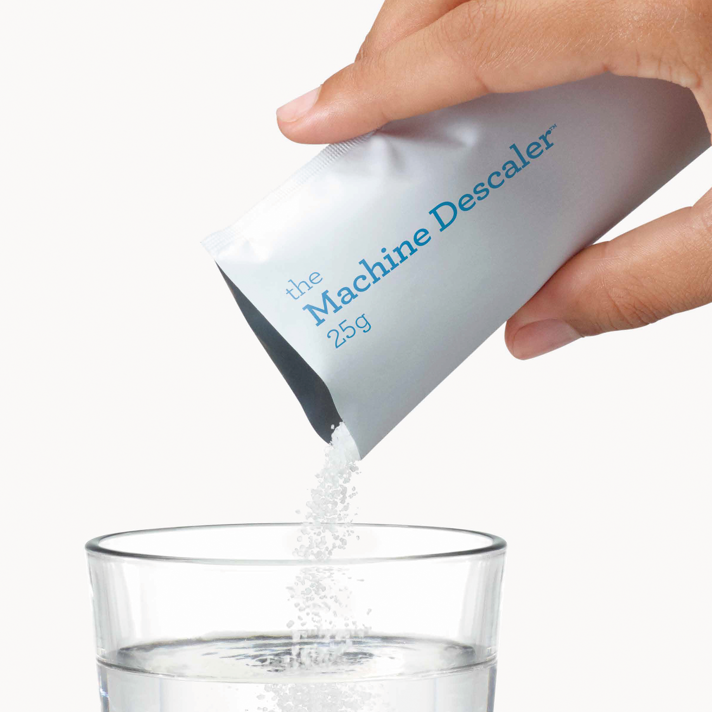 Eine Hand gießt Entkalkerpulver aus einer Packung in ein Glas Wasser. Die Packung ist mit 'the Machine Descaler' beschriftet.