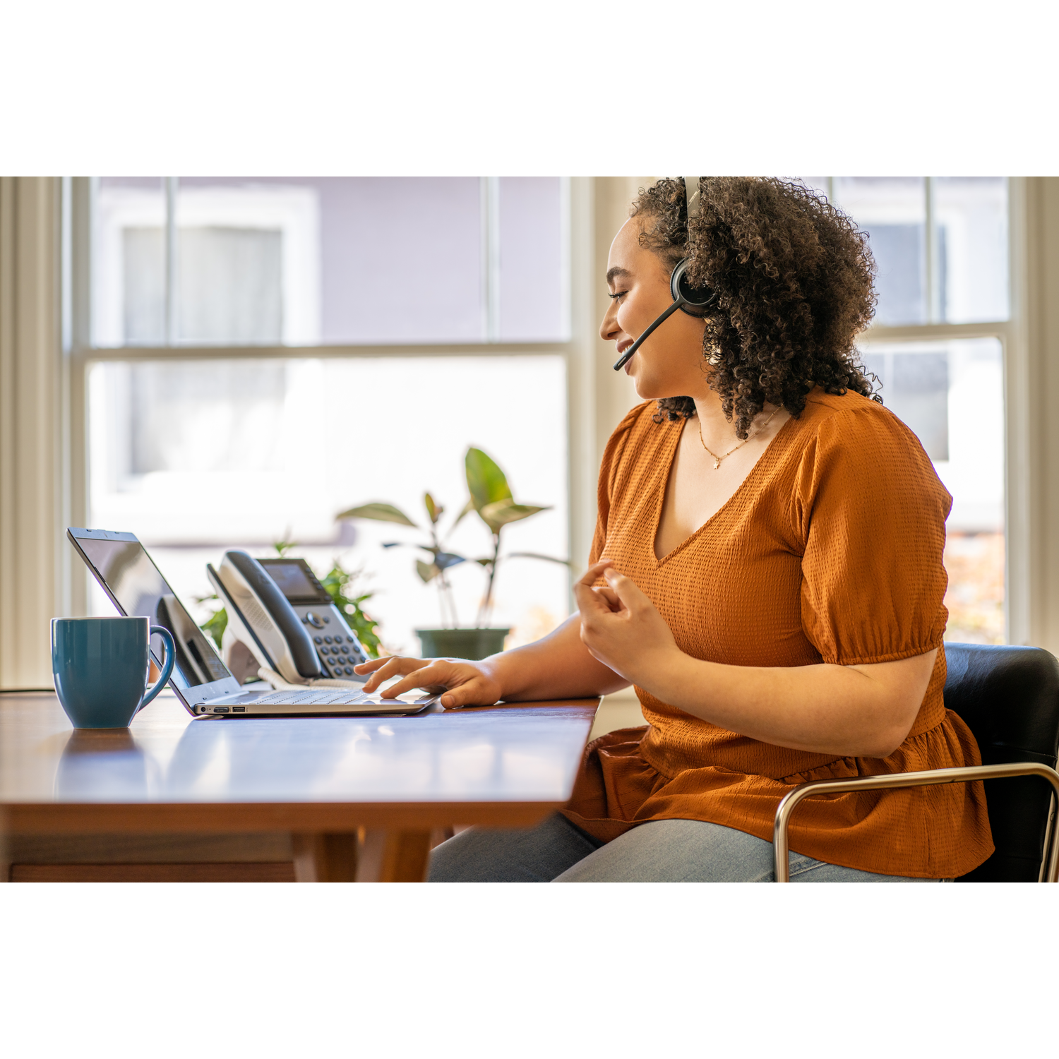 Vrouw met headset werkt op laptop aan bureau. Er is een blauwe mok en telefoon. Ramen op de achtergrond.