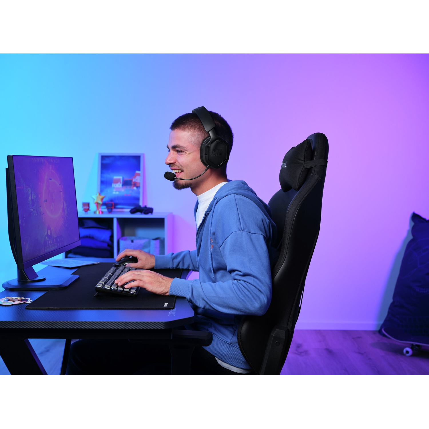 Un uomo con le cuffie sta giocando a una scrivania, usando tastiera e mouse. Un monitor è di fronte a lui. Lo sfondo è viola e blu.