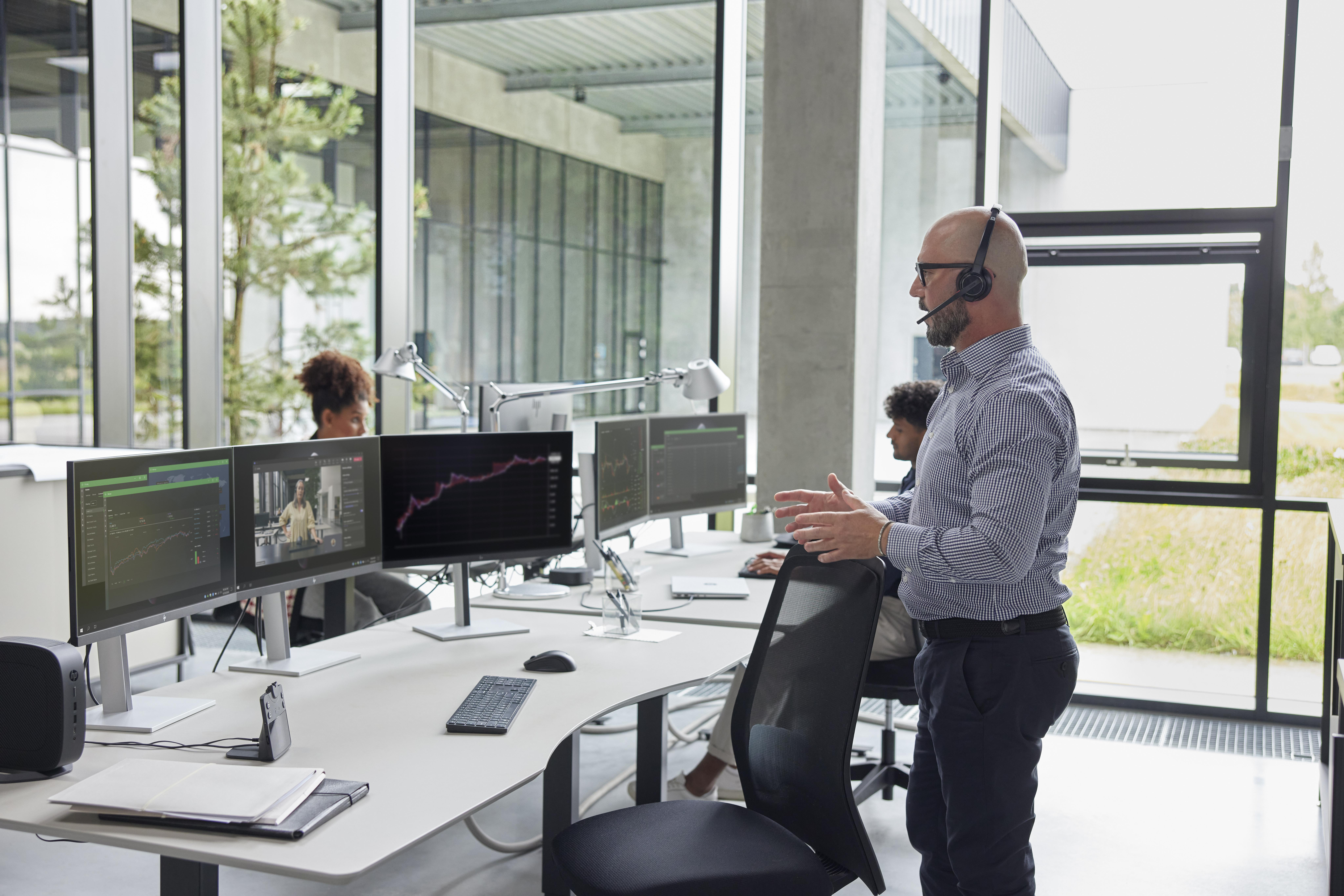 Ein Mann mit Headset gestikuliert in einem Büro. Monitore zeigen Daten und einen Videoanruf. Andere sitzen an Schreibtischen.
