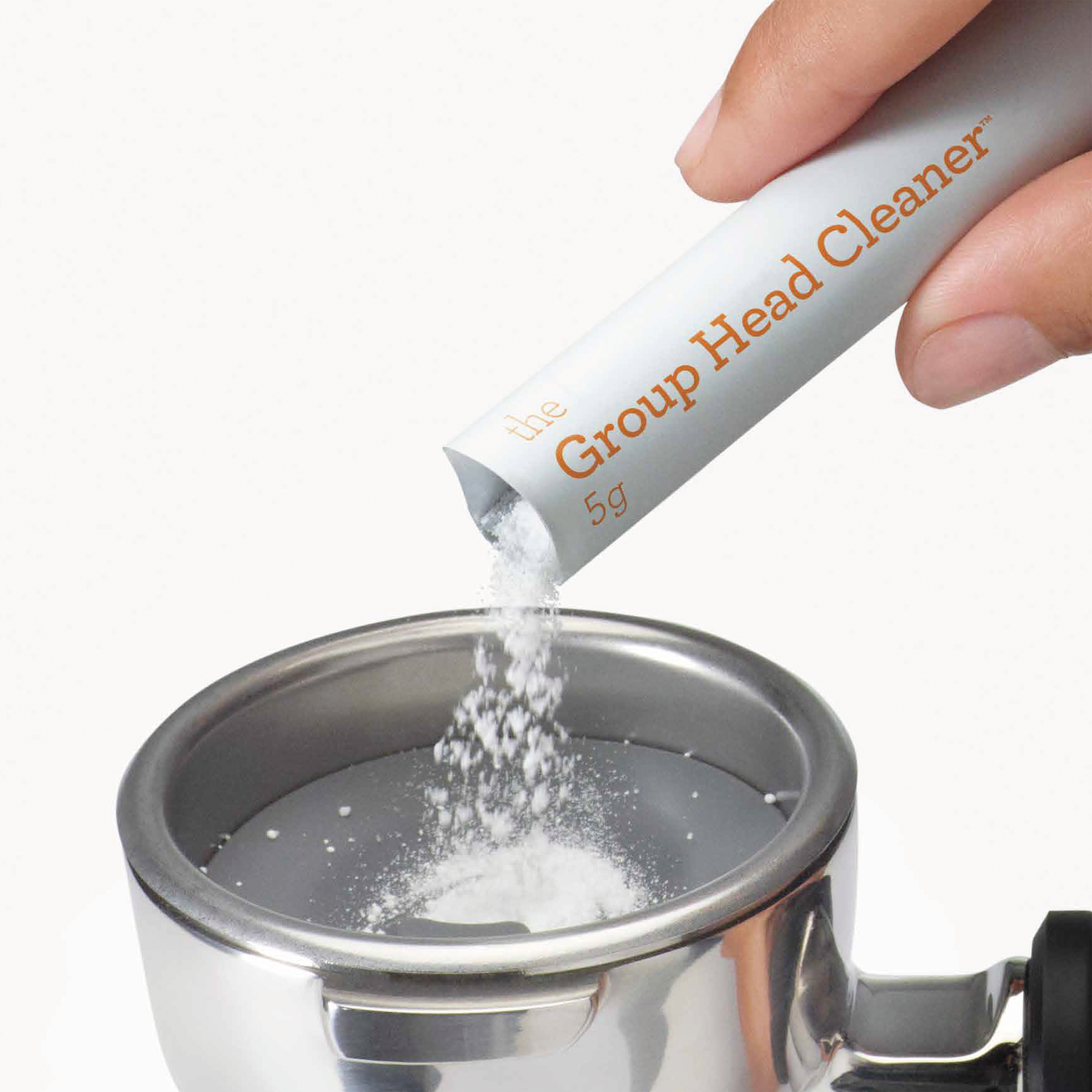 Eine Hand gießt weißes Pulver aus einem Päckchen mit der Aufschrift "Group Head Cleaner" in einen Metall-Siebträger.