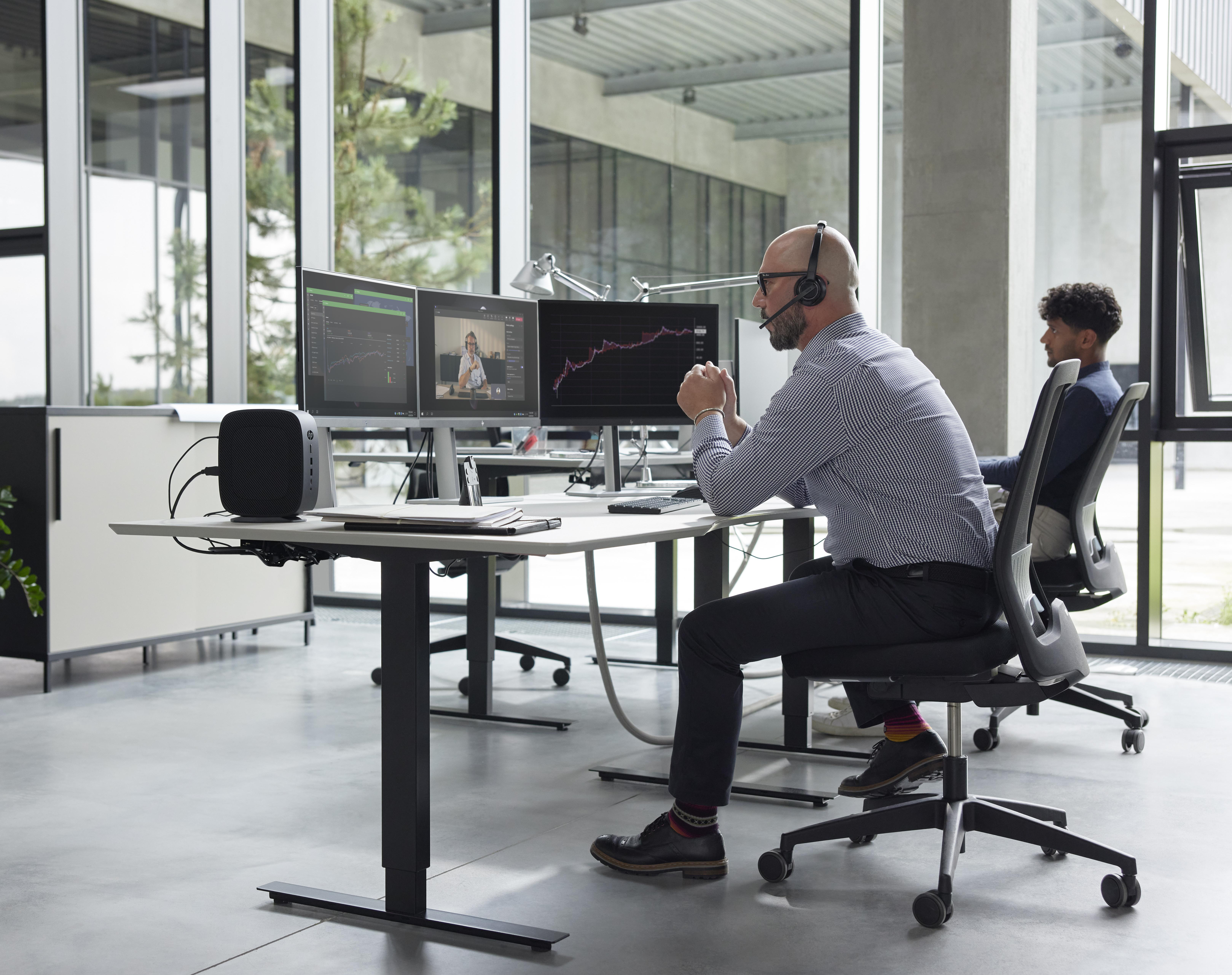 Zwei Männer an Schreibtischen mit Monitoren, die Diagramme und einen Videoanruf anzeigen. Ein Mann trägt ein Headset.