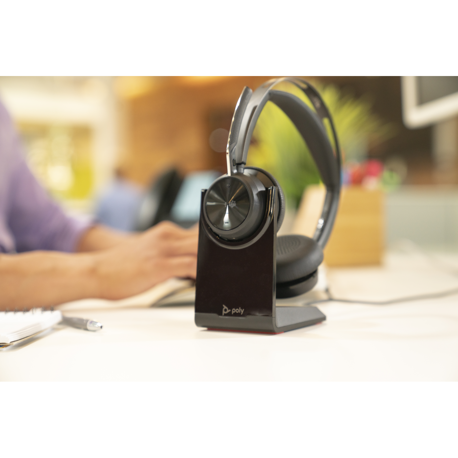 Een zwarte headset op een standaard. Een persoon typt op een laptop op de wazige achtergrond. Een notitieboek en een pen staan op de voorgrond.