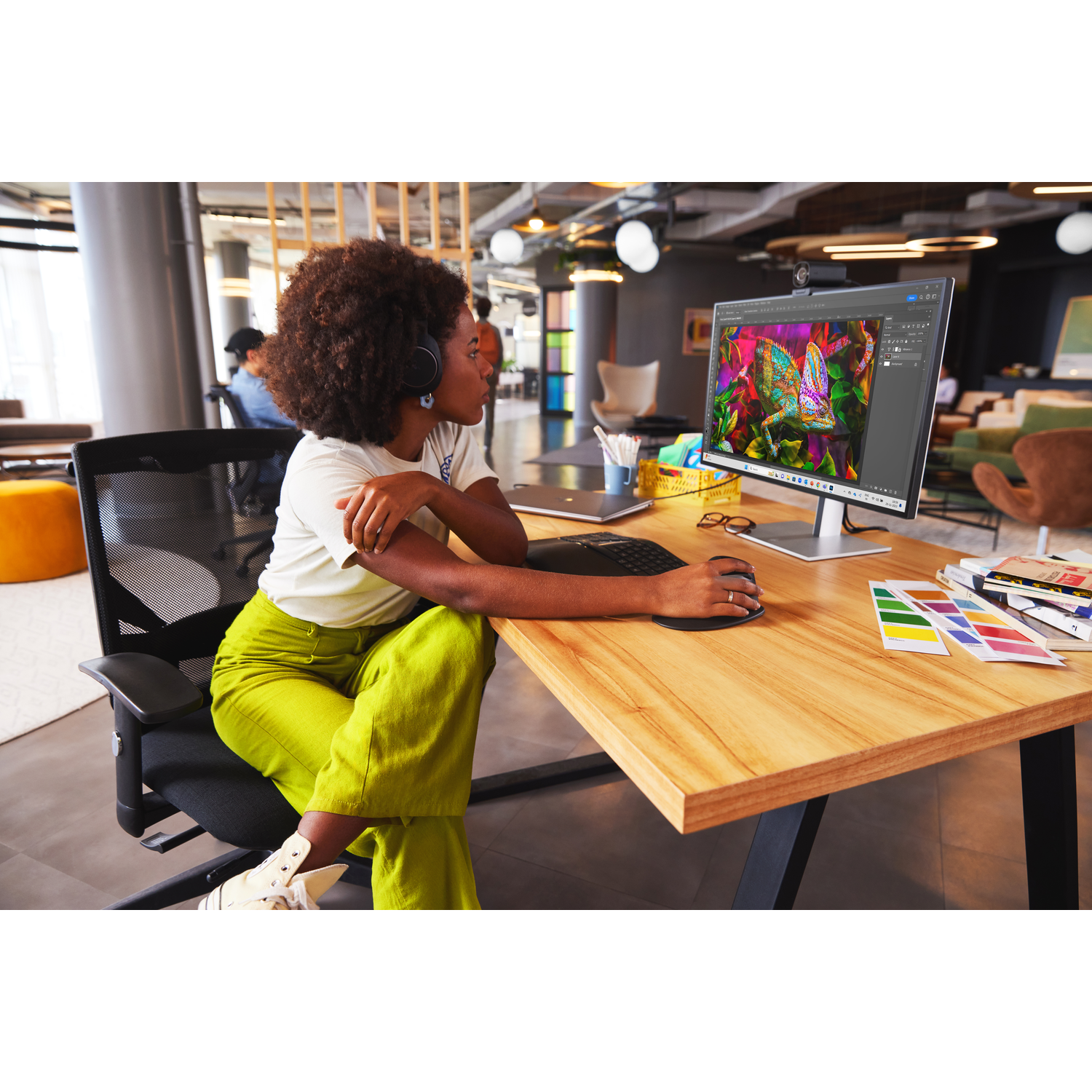 Woman with headphones at desk. Monitor shows photo editing software. Color swatches and books on desk. Other people in background.