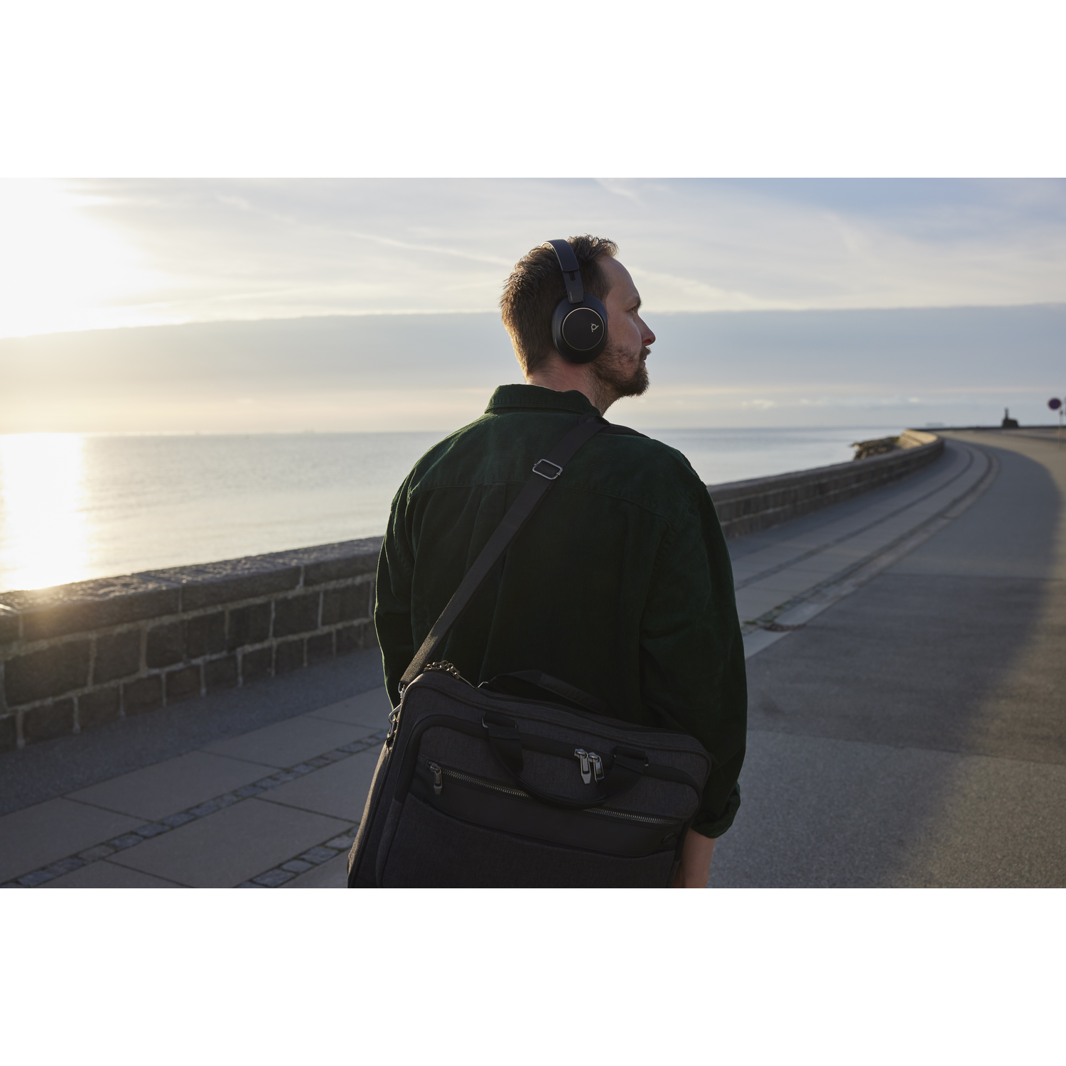 A man wearing headphones and carrying a bag walks along a seaside path. The sea is in the background, with a sunset.