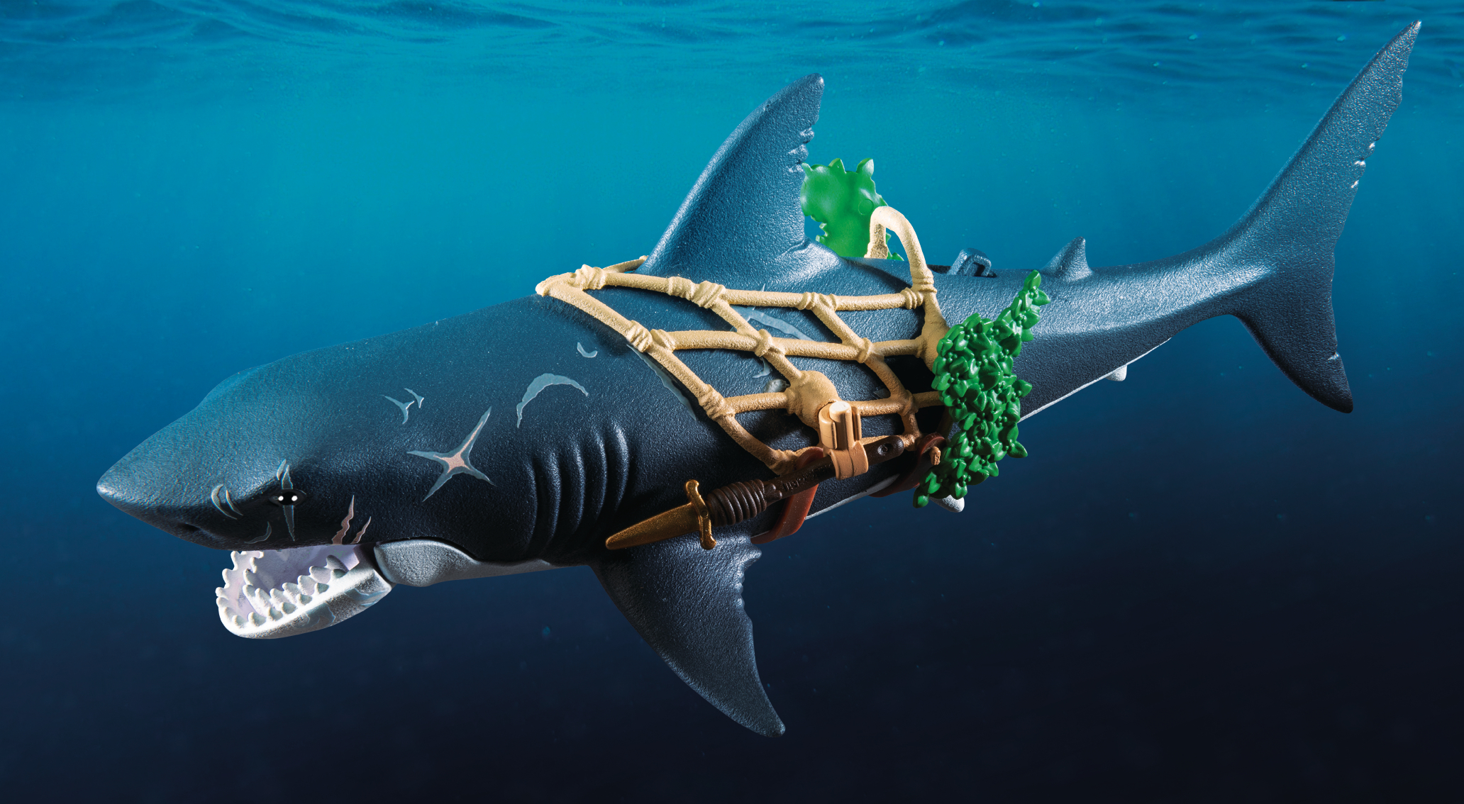 Ein Spielzeug-Hai in blauem Wasser. Es hat ein Geschirr, grünen Seetang und einen Dolch befestigt. Der Hai hat ein geöffnetes Maul und scharfe Zähne.