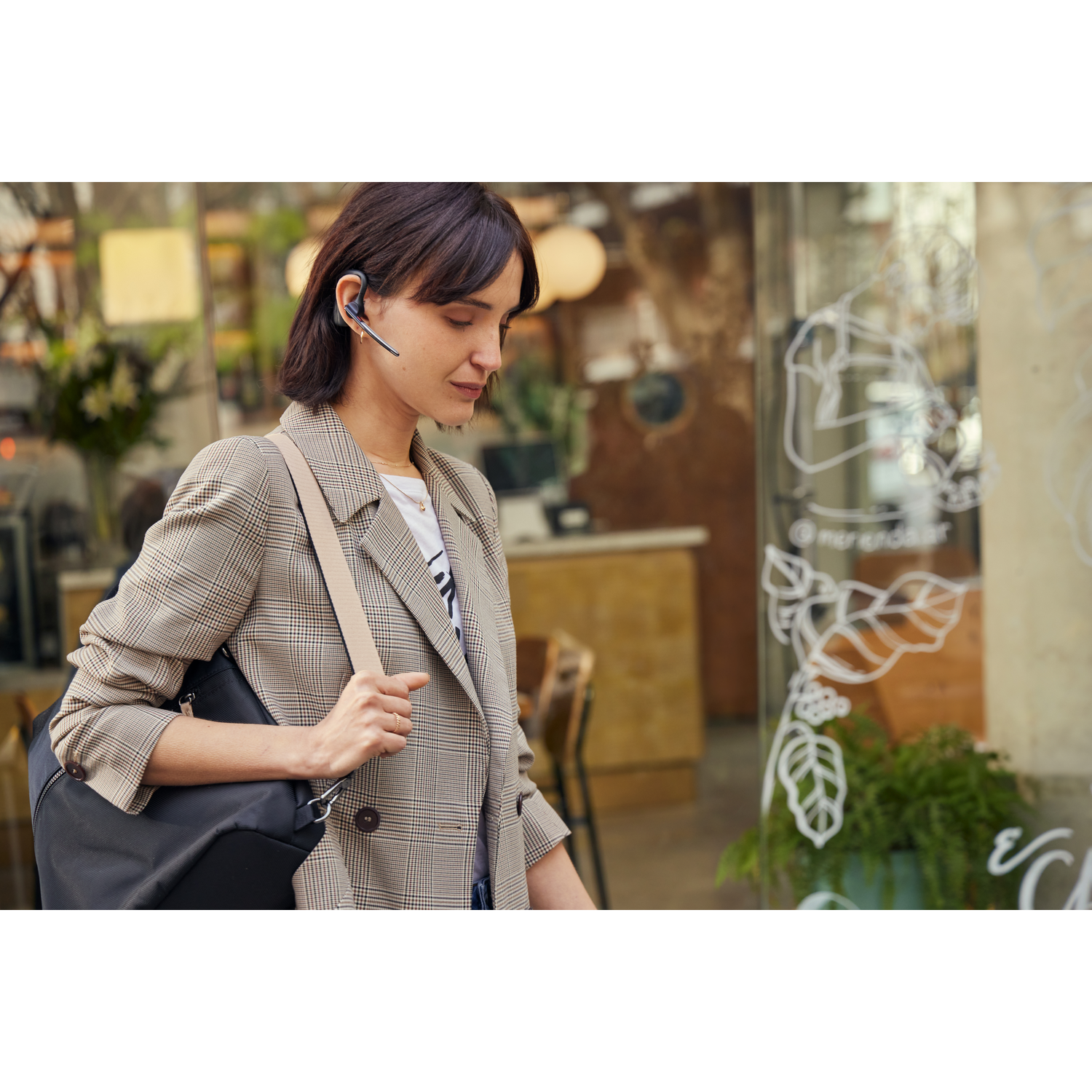 Vrouw met headset en tas loopt langs een caféraam. Ze draagt een geruite blazer en kijkt naar beneden.