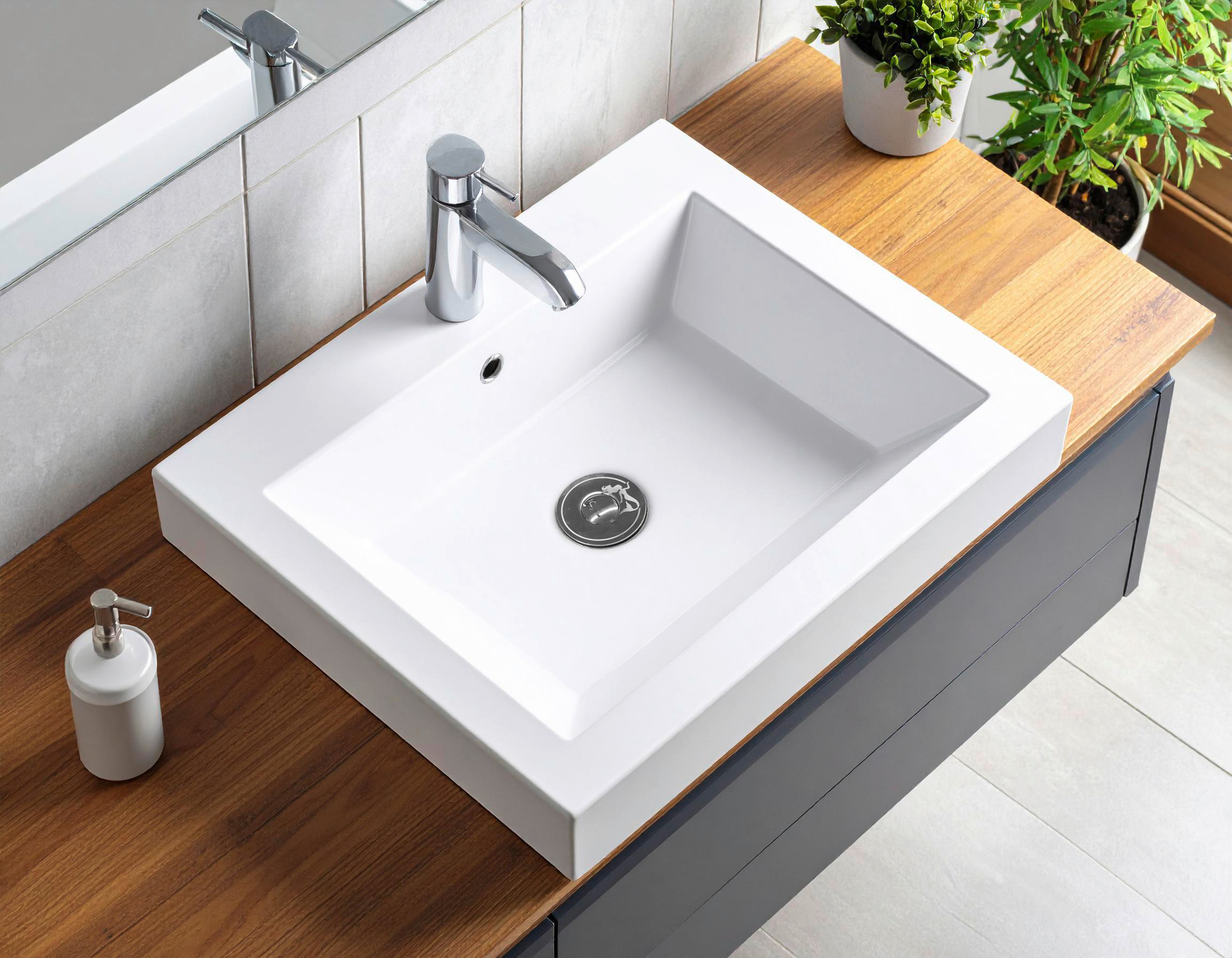 Ein weißes rechteckiges Waschbecken mit einer Chromarmatur steht auf einer Holzplatte. Ein Seifenspender und Pflanzen sind zu sehen.