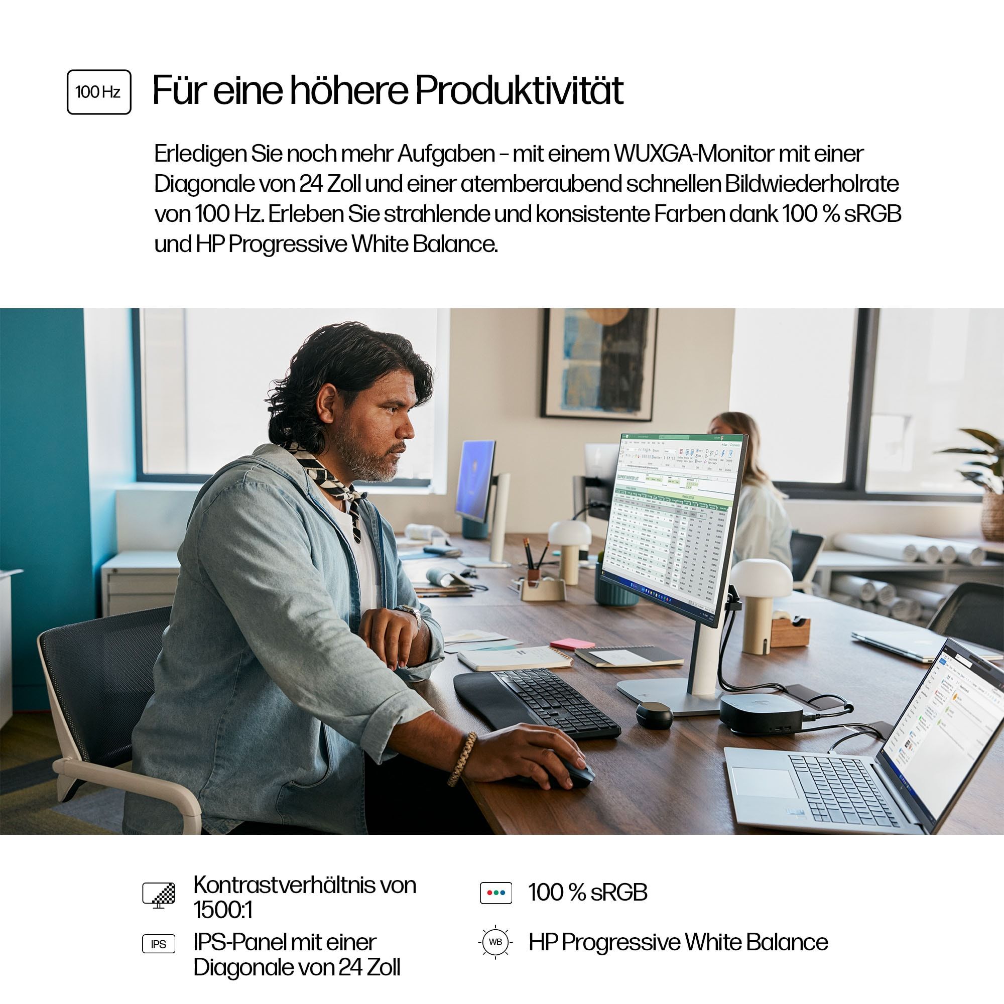 Mann am Schreibtisch benutzt eine Maus; Monitor und Laptop sind geöffnet. Frau im Hintergrund. Der Schreibtisch hat Notizbücher und eine Lampe.