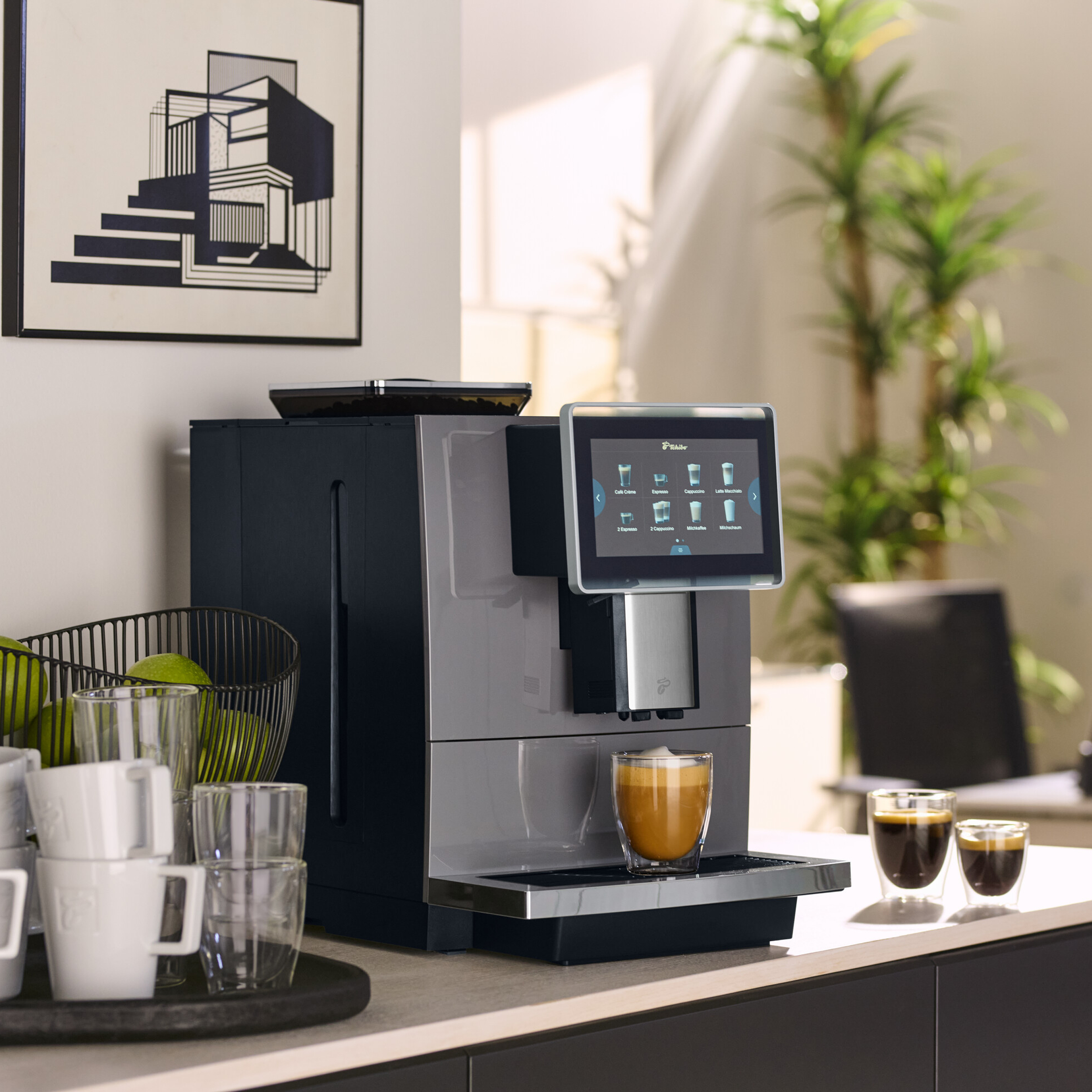 Eine Kaffeemaschine mit Touchscreen zeigt Kaffeeoptionen. Ein Glas Kaffee steht darunter. Äpfel und Tassen sind im Vordergrund.