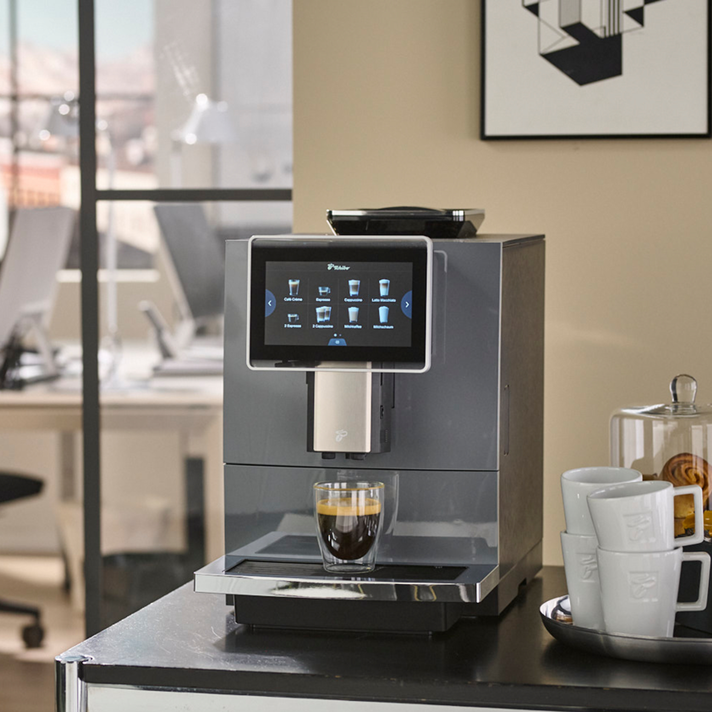 Eine Kaffeemaschine mit Touchscreen, ein Glas Kaffee und weiße Tassen auf einem Tisch.