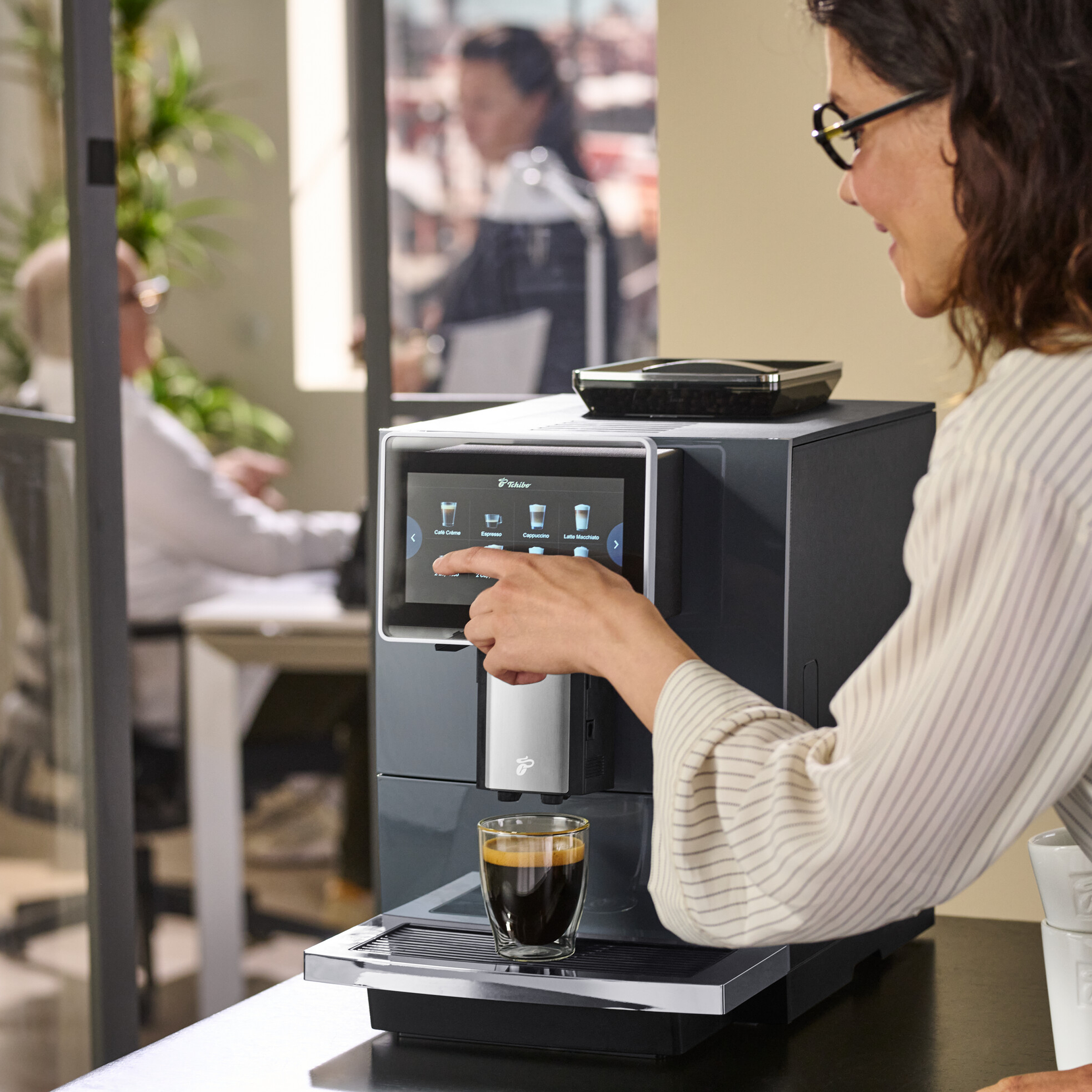 Frau bedient Kaffeemaschine. Sie berührt den Bildschirm. Ein Glas Espresso ist fertig. Andere Leute arbeiten im Hintergrund.