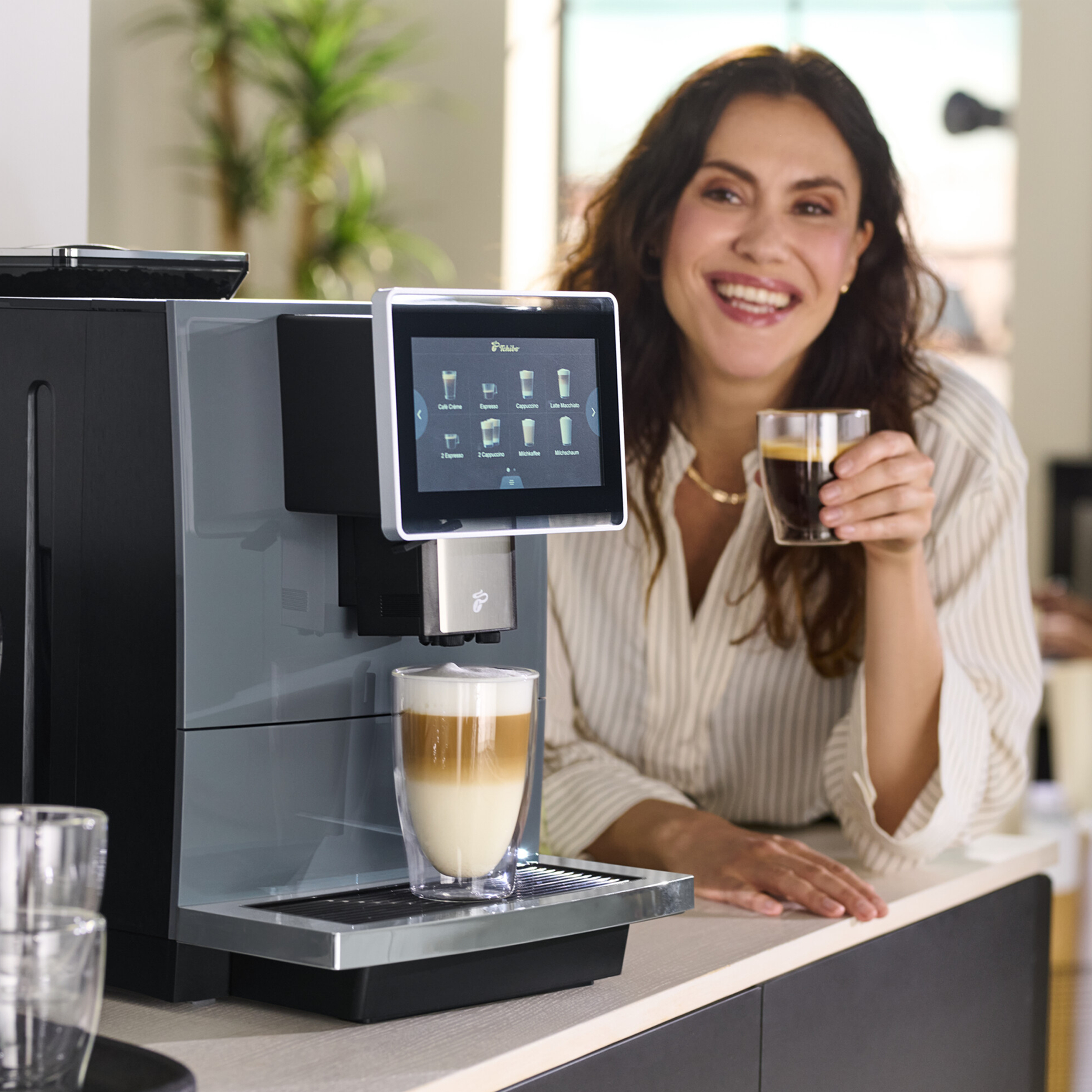 Eine lächelnde Frau hält Kaffee in der Nähe einer Kaffeemaschine. Ein Glas Latte steht davor. Der Hintergrund hat Pflanzen.