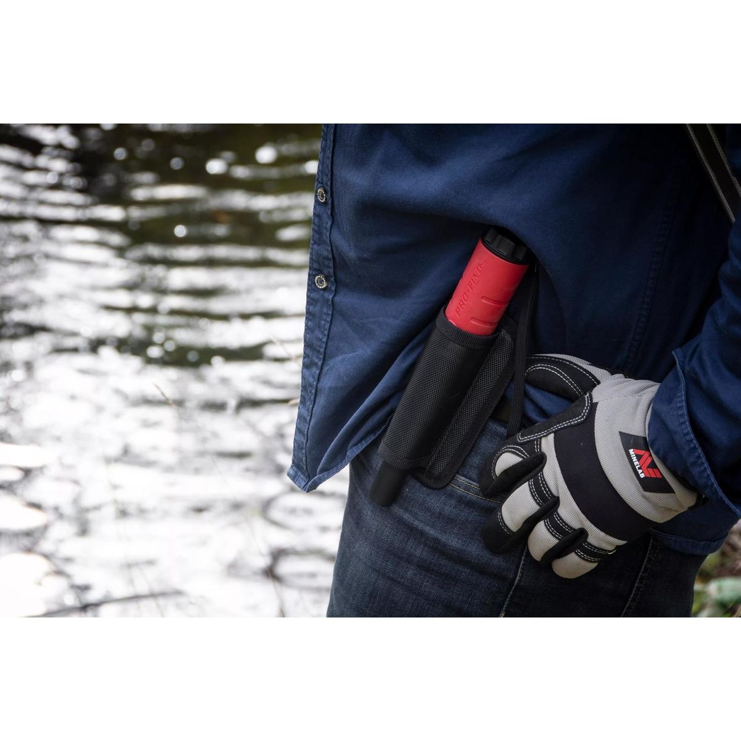 Una persona indossa una camicia blu e jeans, tiene un metal detector e indossa un guanto. Un fiume è sullo sfondo.