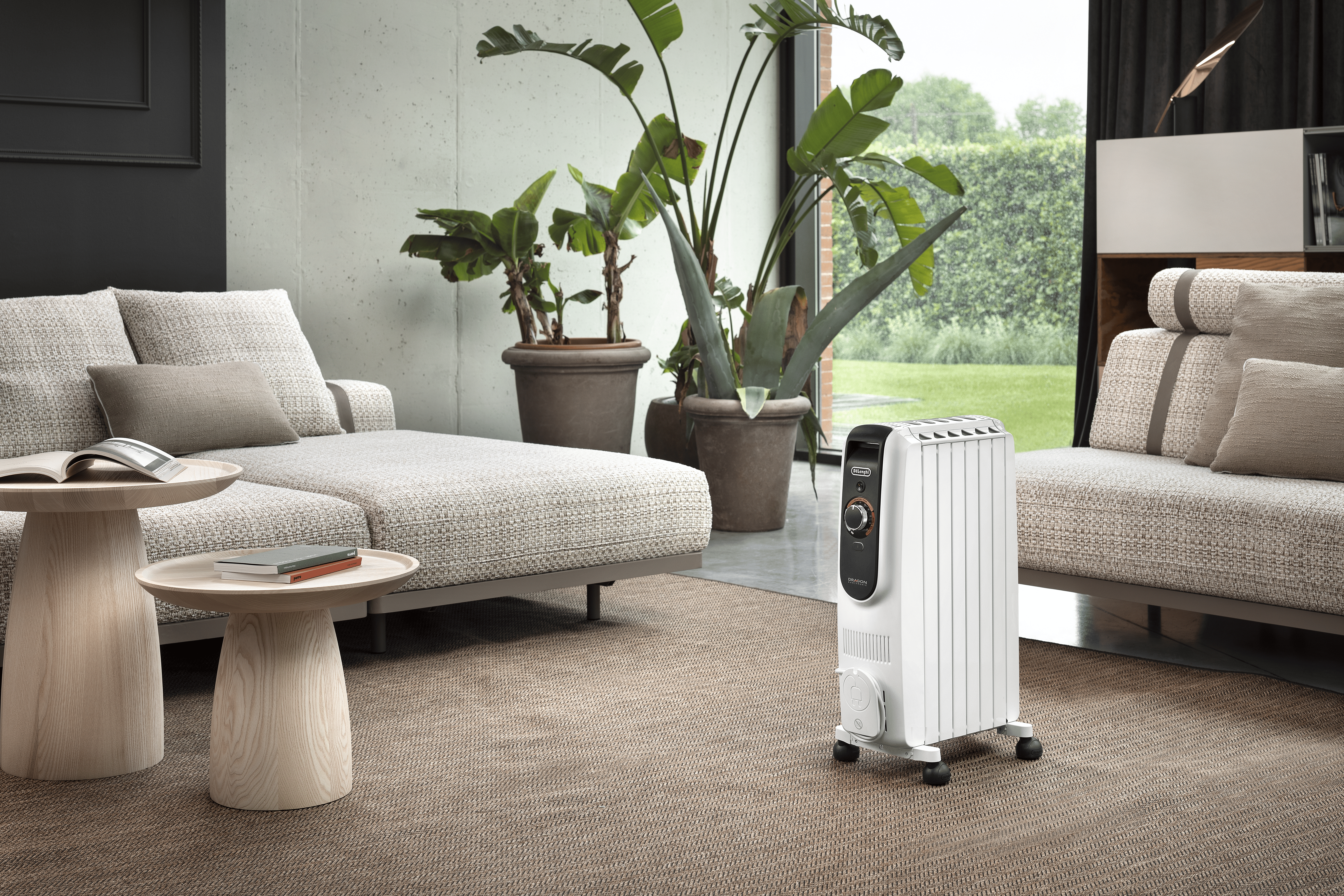 Modernes Wohnzimmer mit Sofas, Pflanzen und weißem Ölradiator. Draußen ist ein grüner Garten durch ein Fenster sichtbar.