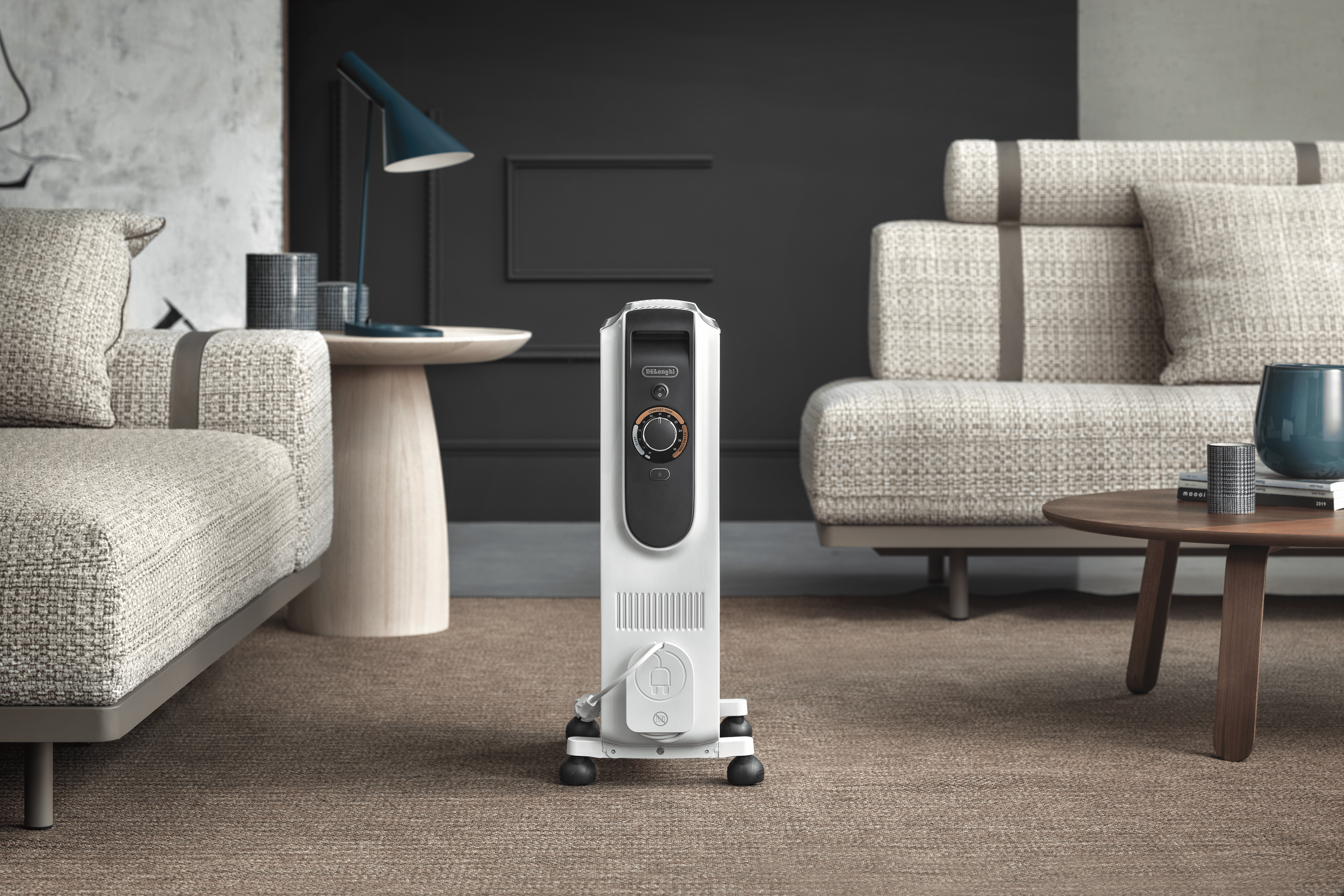 Ein weißer elektrischer Heizkörper steht in einem Wohnzimmer. Sofas und ein Tisch sind vorhanden.