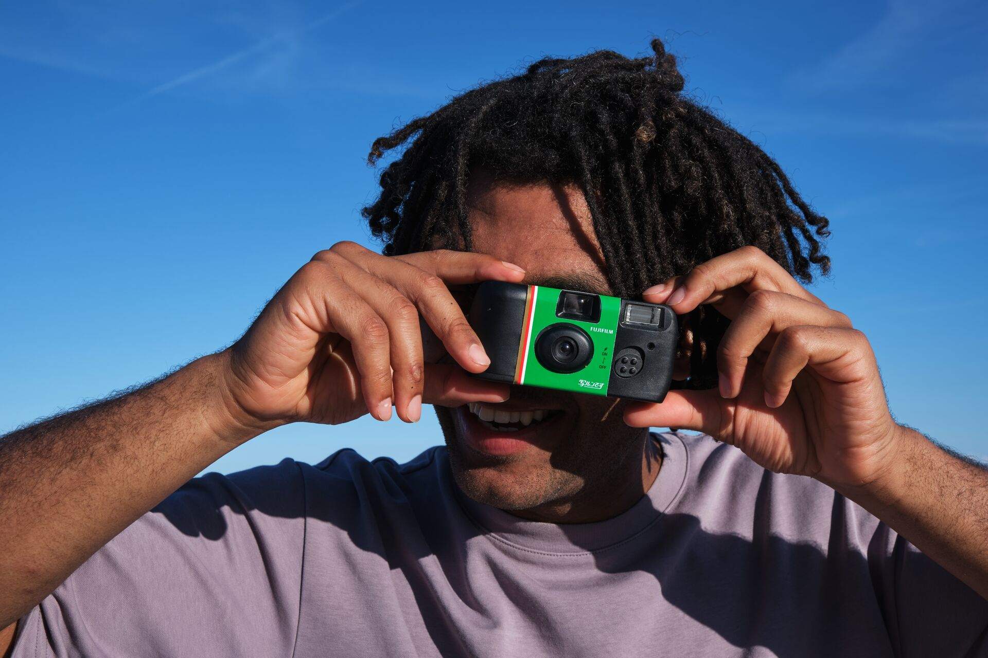 Eine Person hält eine Fujifilm-Kamera und lächelt. Die Kamera hat grüne und rote Streifen. Klarer blauer Himmel im Hintergrund.