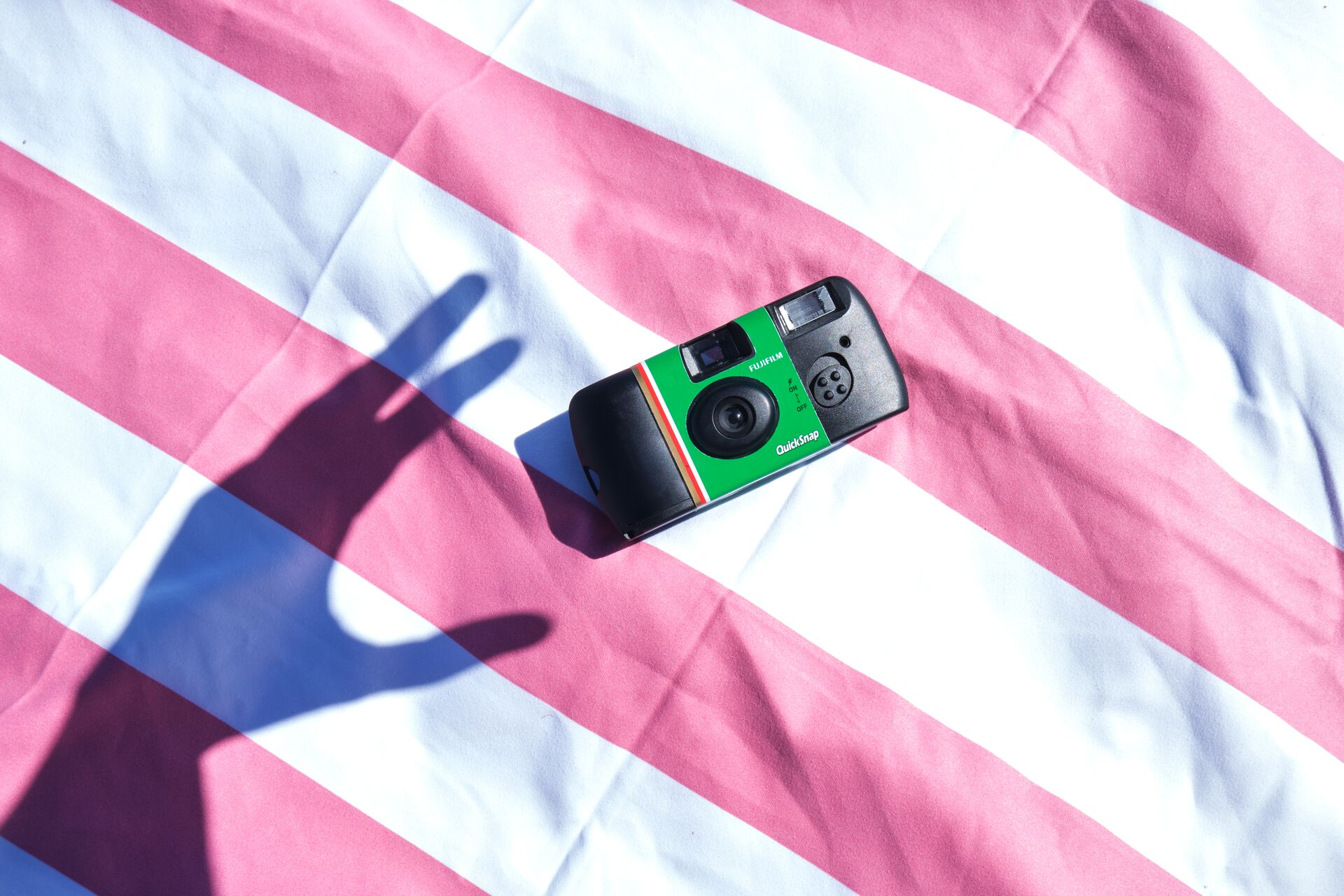 Eine schwarze und grüne Fujifilm QuickSnap Kamera liegt auf einer rosa-weiß gestreiften Oberfläche. Ein Handschatten wird geworfen.