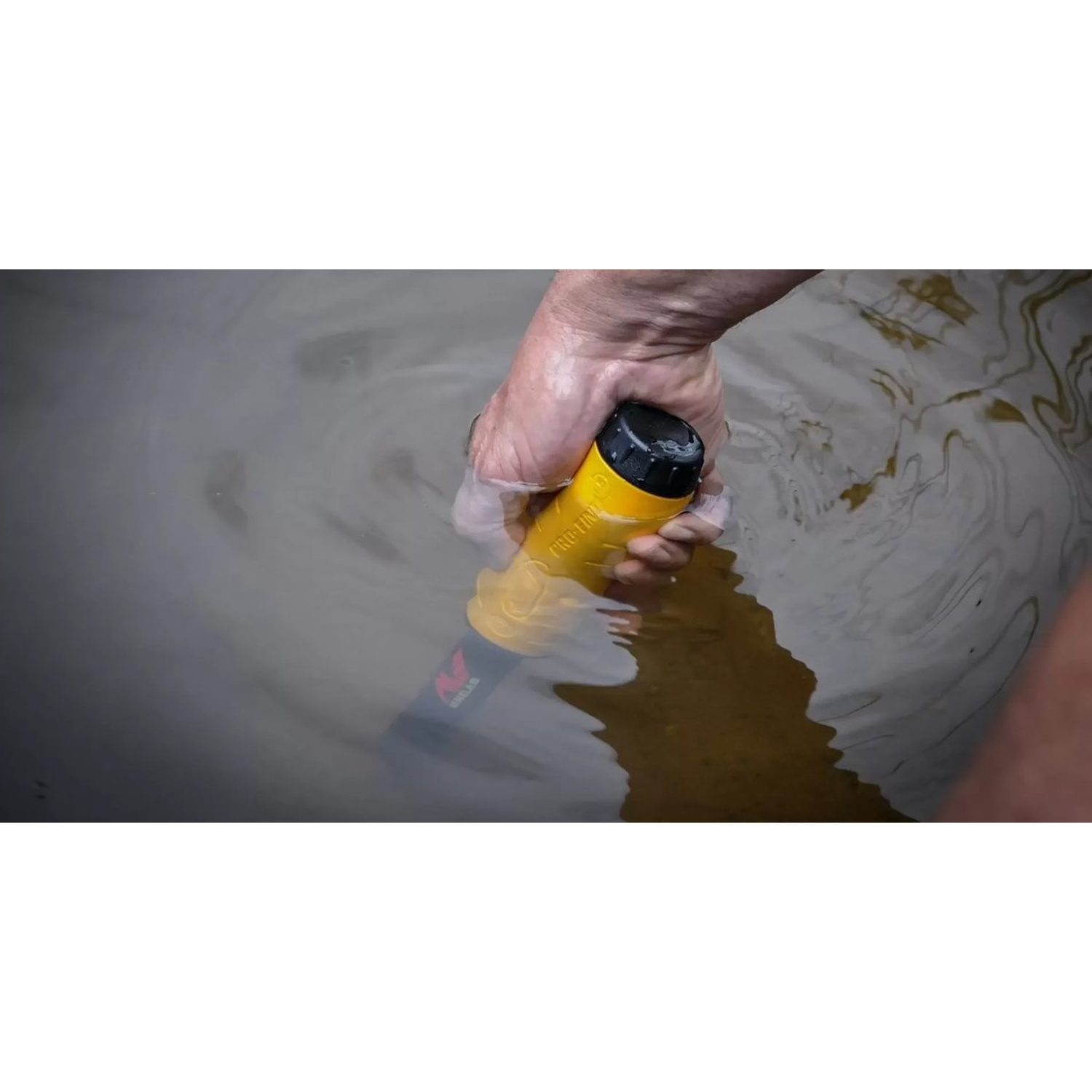 Una mano tiene un metal detector giallo sott'acqua. Il dispositivo è parzialmente immerso in acqua torbida.