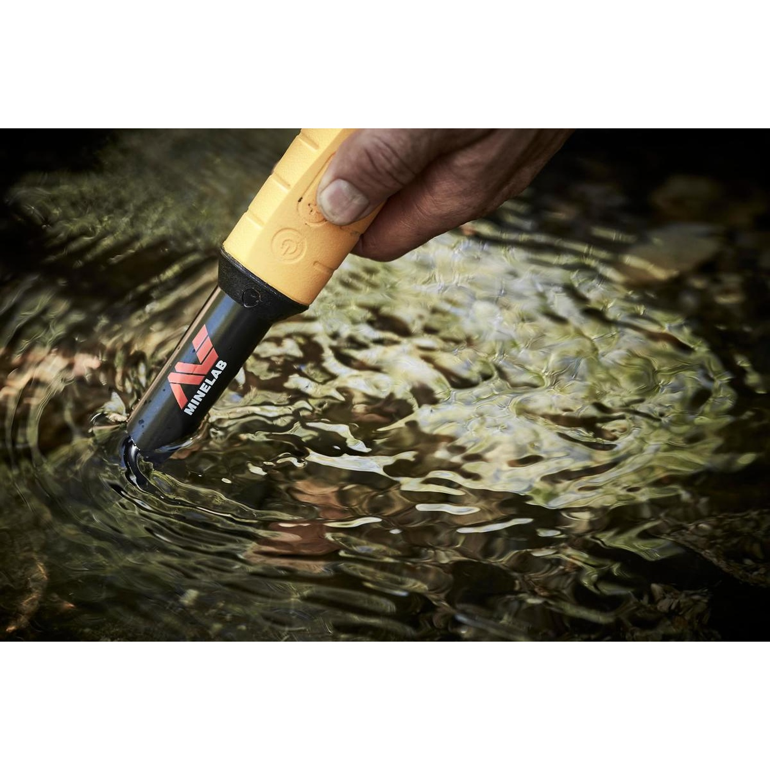 Una mano tiene un metal detector giallo nell'acqua. La punta del metal detector è immersa. Increspature e riflessi sono visibili sulla superficie dell'acqua.