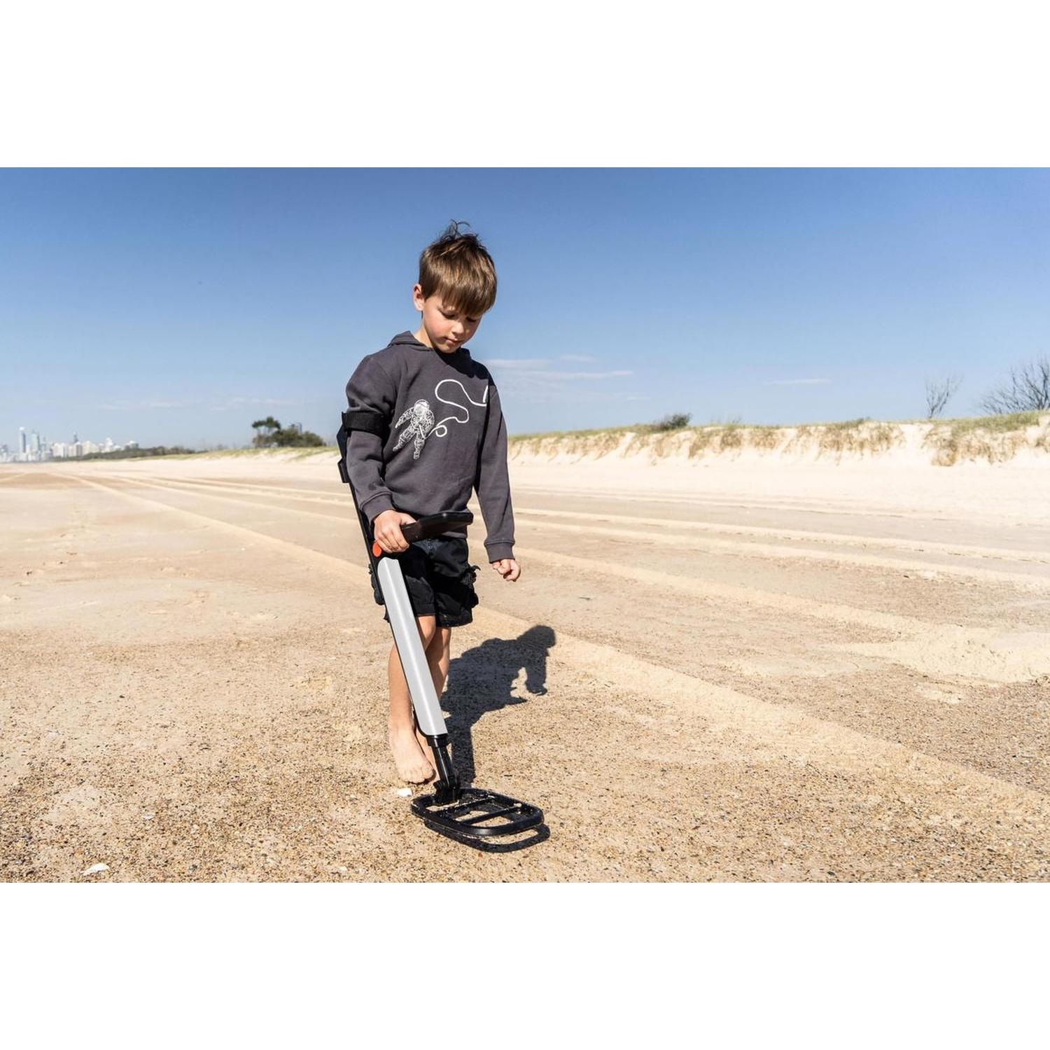 Un ragazzo sulla spiaggia usa un metal detector. Indossa una felpa grigia e pantaloncini neri. Il cielo è blu e la sabbia è beige.