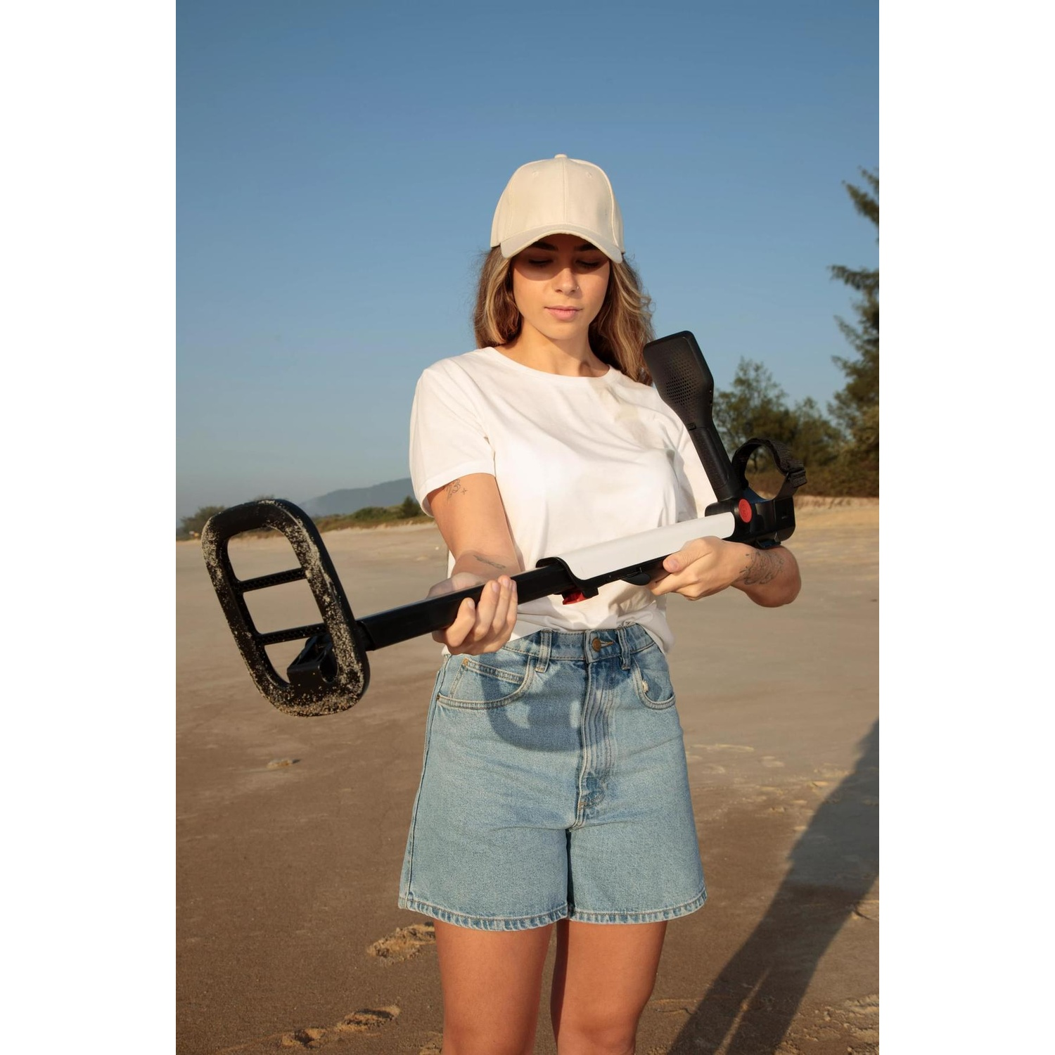 Donna sulla spiaggia con un metal detector. Indossa un berretto, una maglietta bianca e pantaloncini di jeans. Sabbia e mare sullo sfondo.