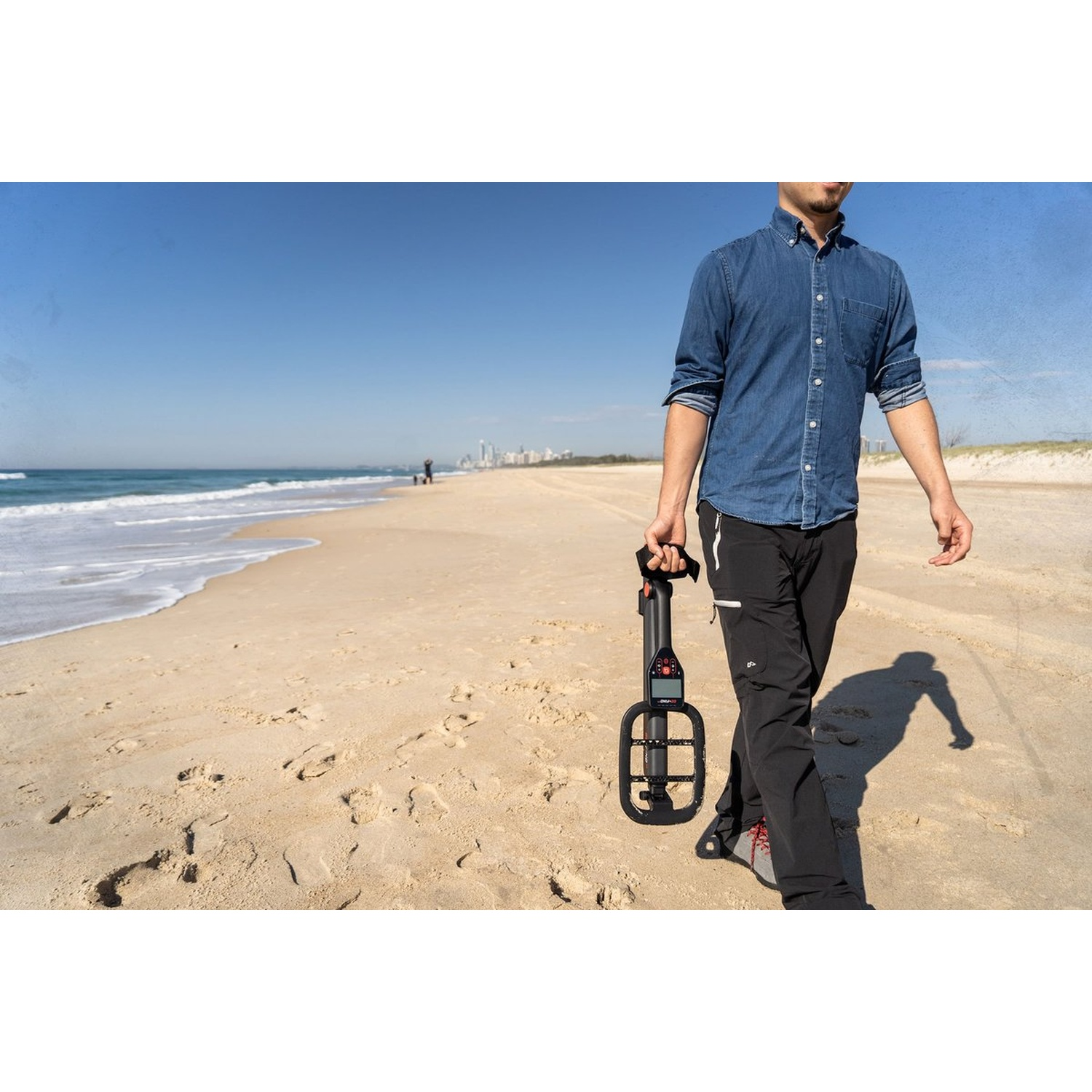 Uomo cammina sulla spiaggia con metal detector. Cielo blu, sabbia e oceano sullo sfondo.