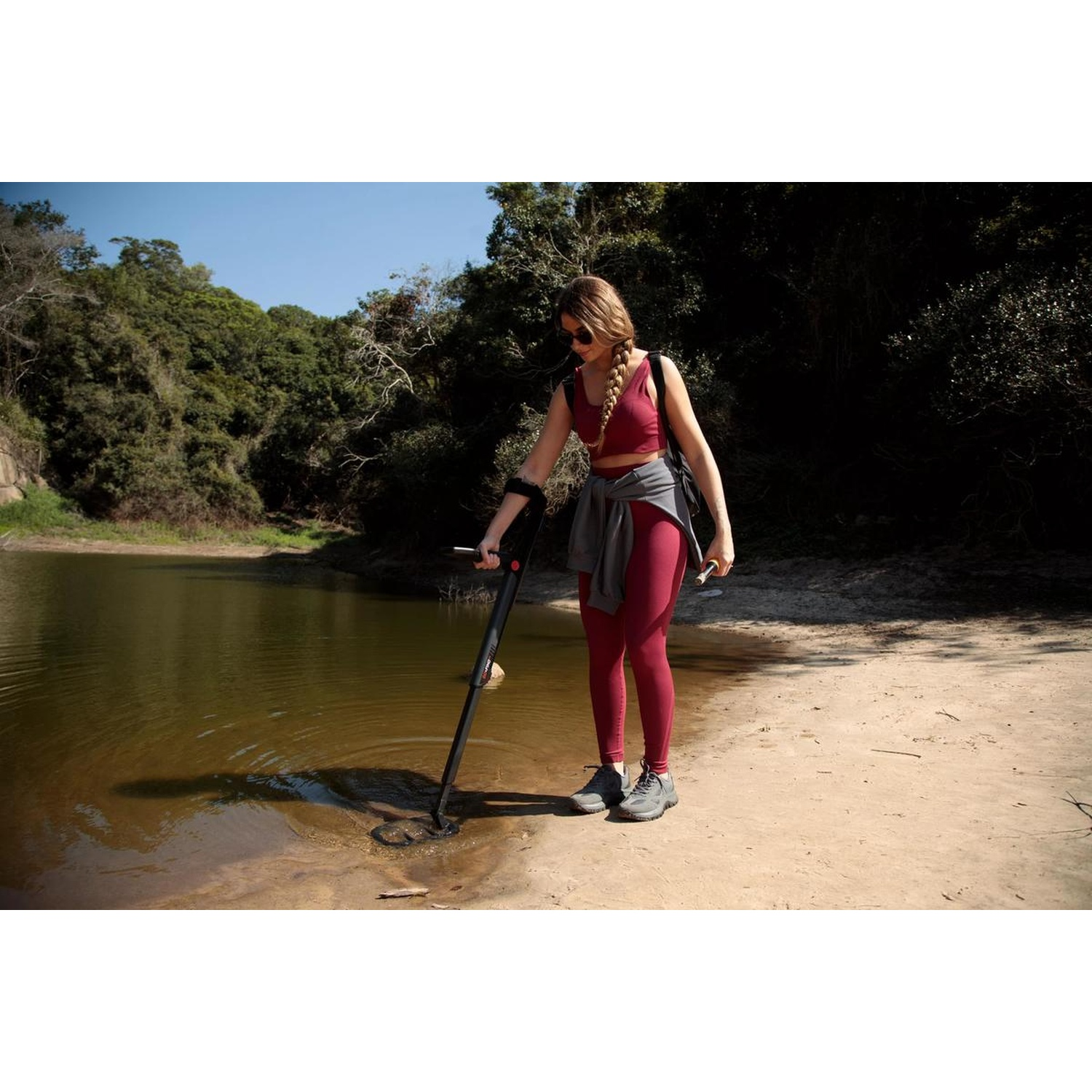 Una donna usa un metal detector sulla riva di un lago. Indossa abbigliamento sportivo rosso, occhiali da sole e tiene un piccolo oggetto.
