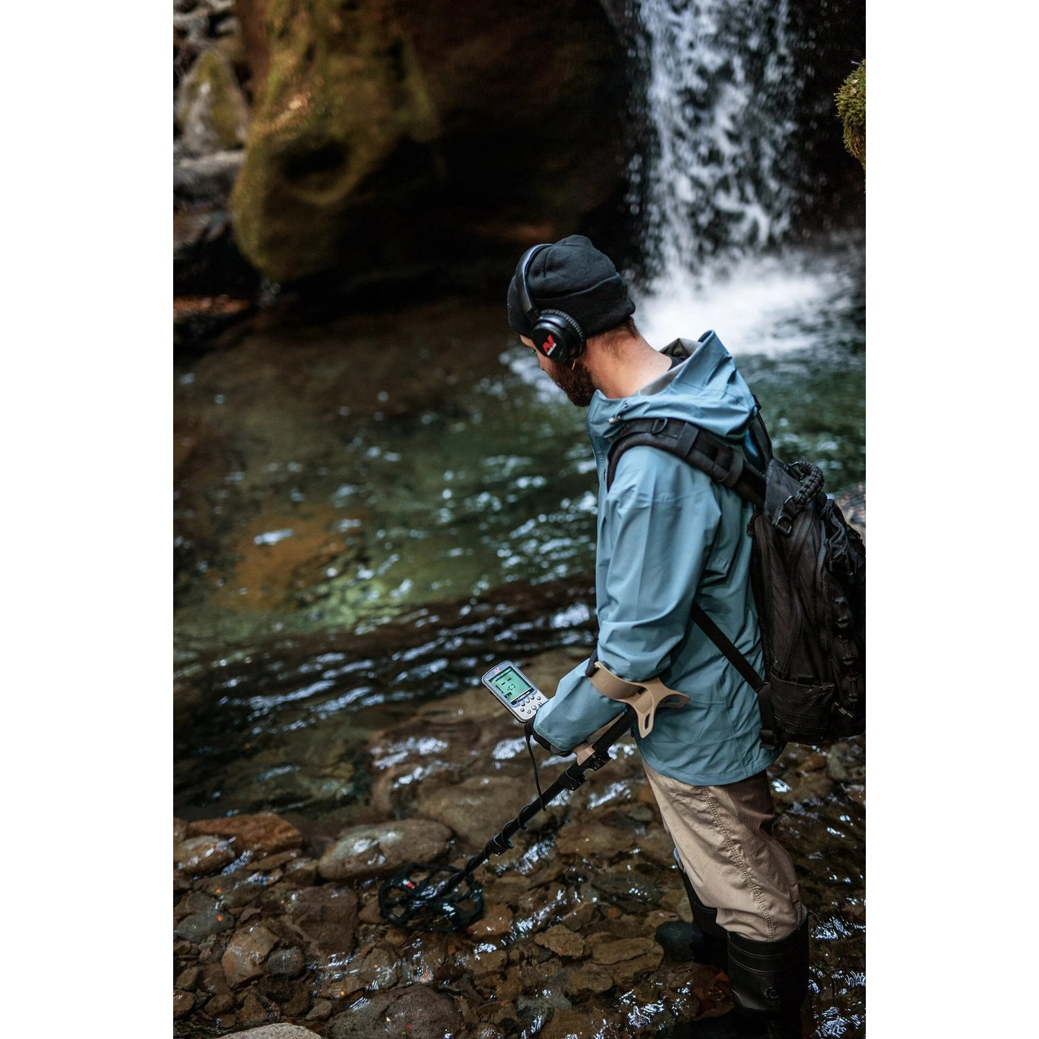 Una persona in un torrente usa un metal detector vicino a una cascata. Indossa cuffie, una giacca e stivali. Lo sfondo è roccioso.