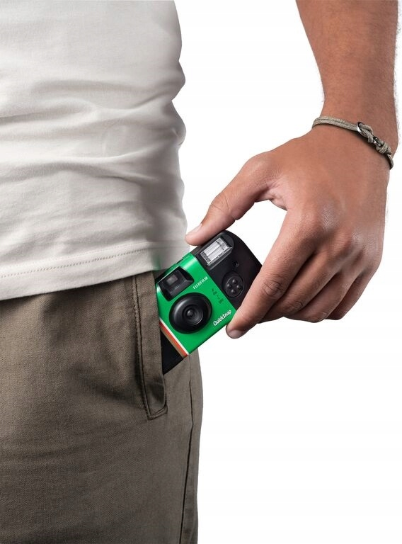 Ręka osoby trzyma zielony aparat z kieszeni. Aparat to Fujifilm QuickSnap. Osoba ma na sobie bransoletkę i białą koszulę.