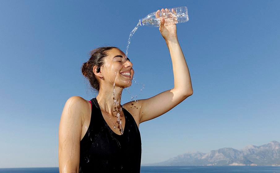 Frau gießt sich Wasser aus einer Flasche ins Gesicht. Sie lächelt mit geschlossenen Augen. Blauer Himmel, Meer und Berge im Hintergrund.