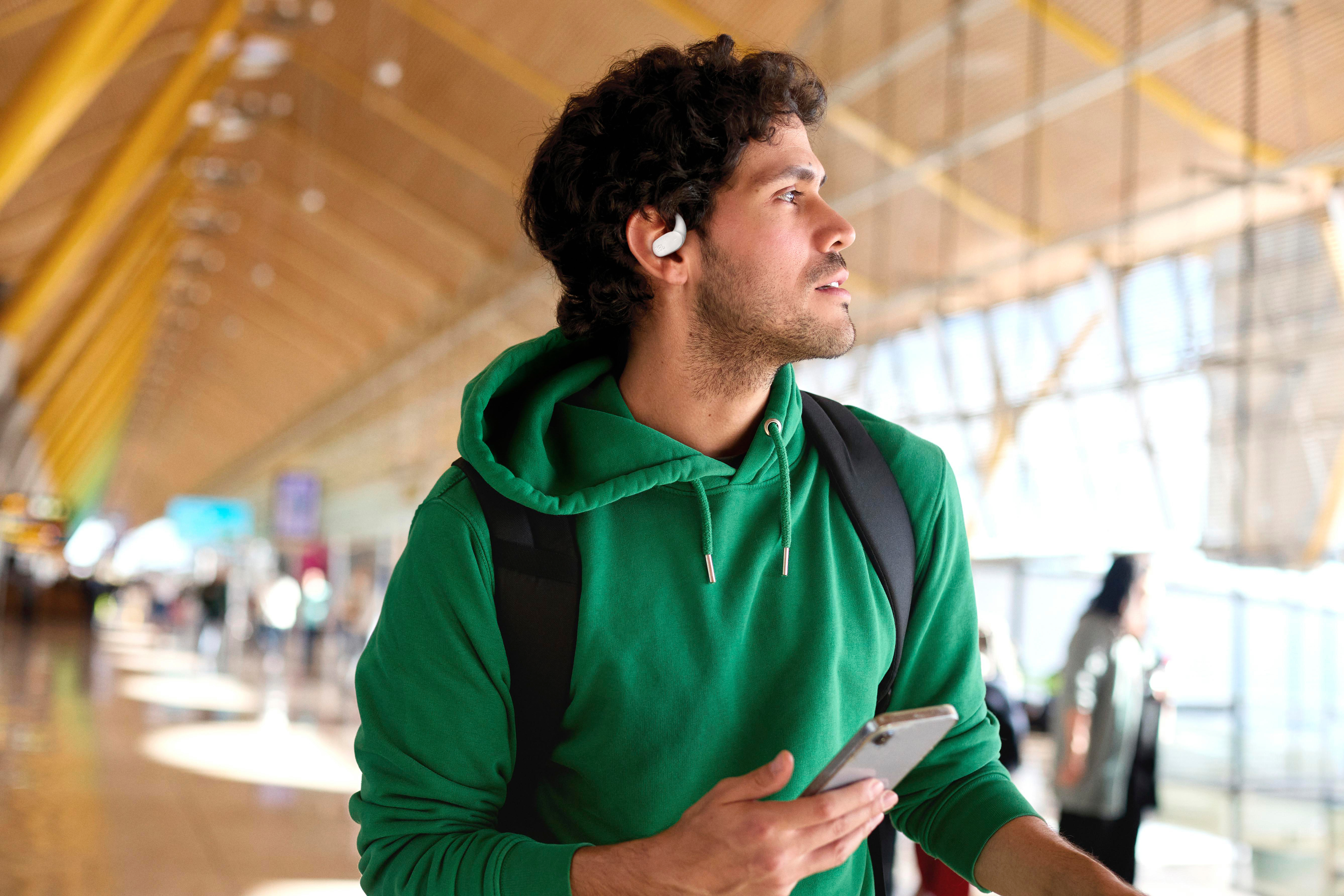 Mann in grünem Kapuzenpullover mit Ohrhörer und Telefon am Flughafen. Hintergrund hat Fenster und Menschen.