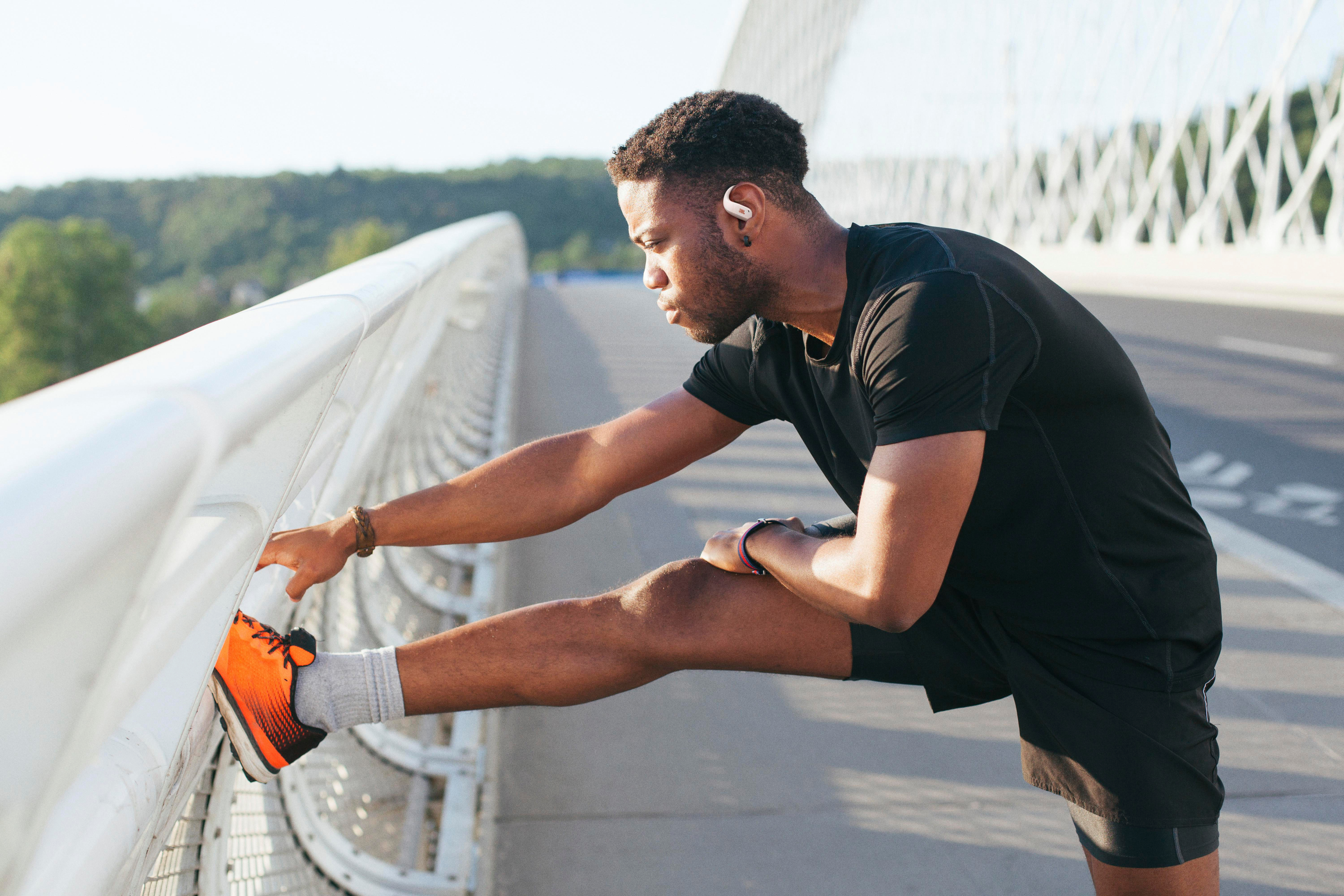 Ein Mann dehnt sein Bein auf einer Brücke, trägt schwarze Sportkleidung. Er hat Ohrhörer und orangefarbene Schuhe.