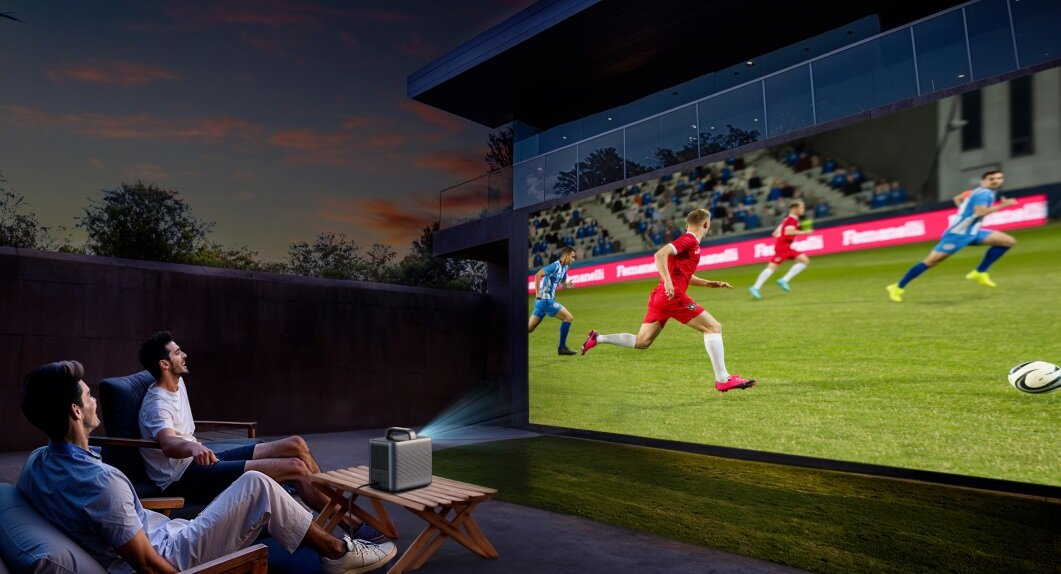 Mężczyźni oglądają mecz piłki nożnej wyświetlany na ekranie. Projektor jest na stole. Gracze biegają po zielonej trawie.