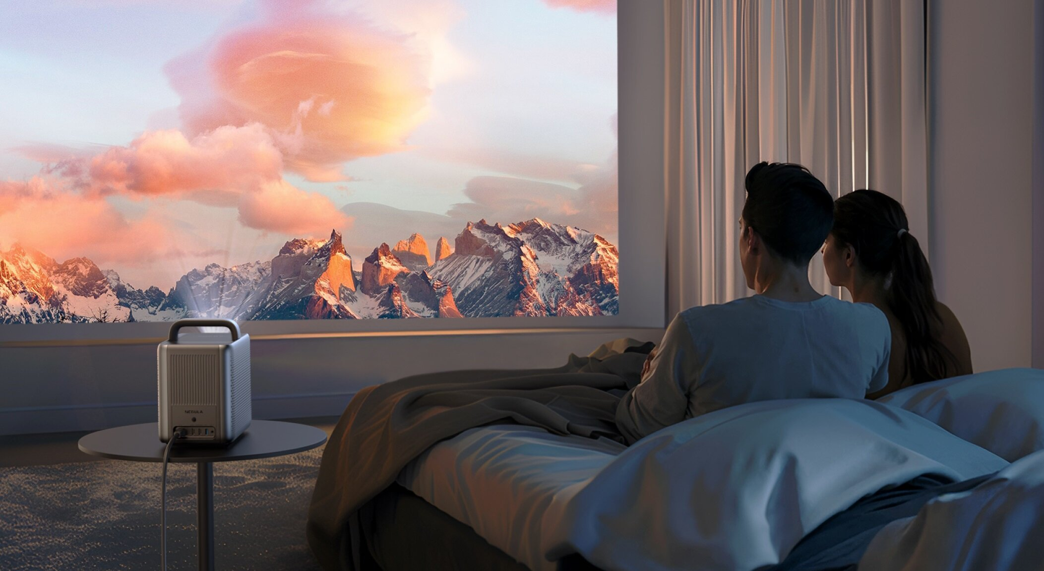 Para ogląda górski krajobraz wyświetlany na ścianie. Projektor stoi na stoliku obok łóżka.