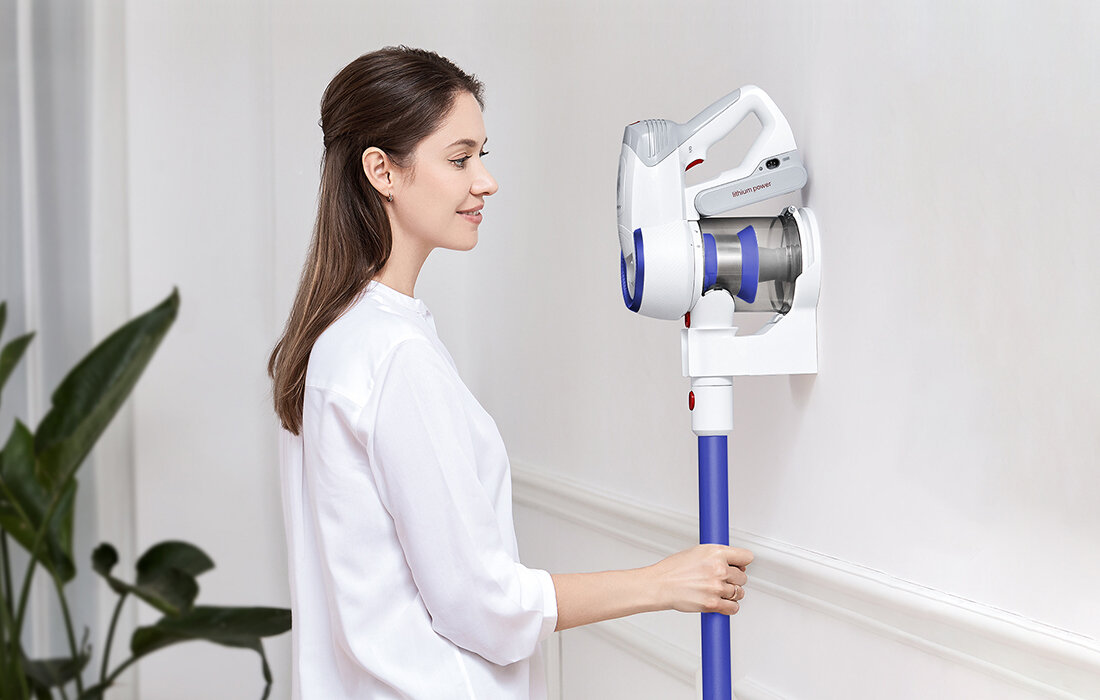 Kobieta w białej koszuli trzyma niebiesko-biały odkurzacz zamontowany na ścianie. Uśmiecha się do odkurzacza.