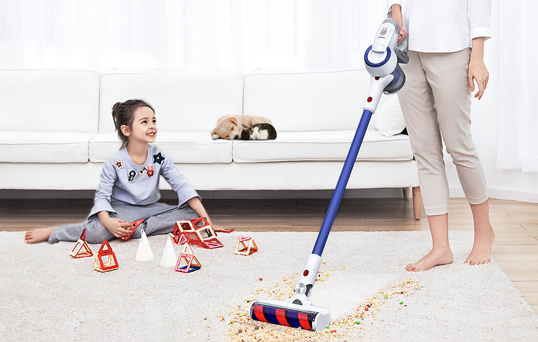 Dziewczynka siedzi na podłodze bawiąc się zabawkami, obok odkurzacza. Kobieta odkurza dywan. Pies śpi na kanapie.