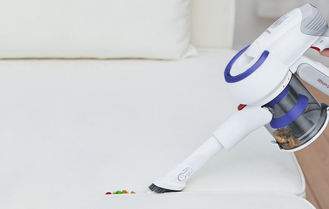 Biały odkurzacz czyści białe łóżko. Na łóżku znajdują się resztki, w tym czerwone i zielone kawałki. Poduszka w tle.