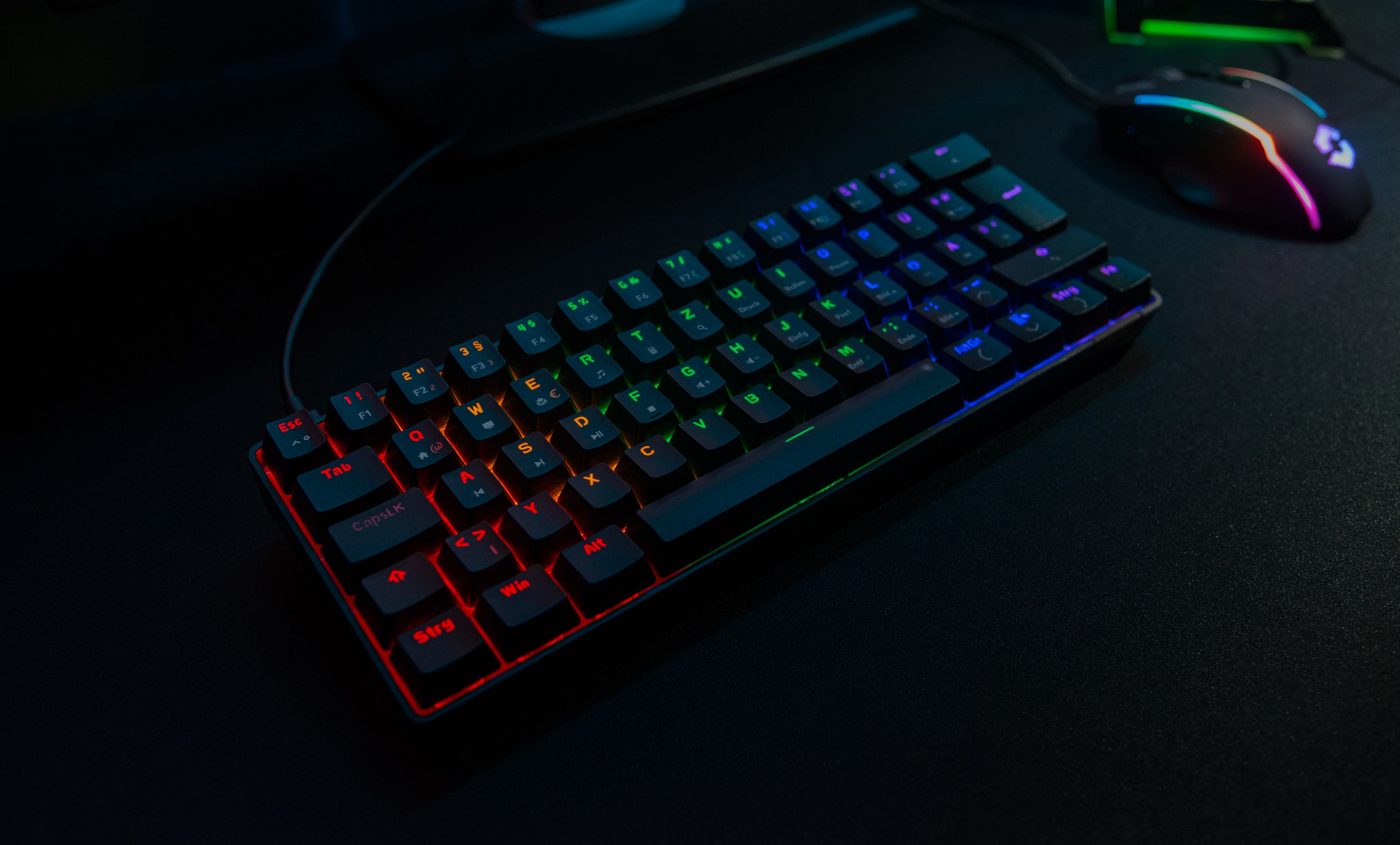Eine Gaming-Tastatur mit Regenbogenlichtern und eine Maus auf dunkler Oberfläche. Tasten leuchten.