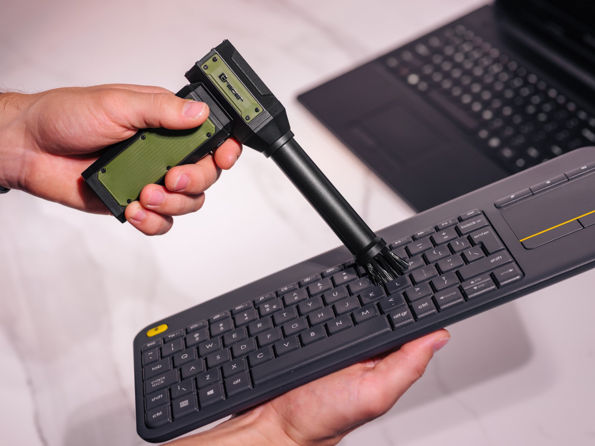 Osoba czyści klawiaturę za pomocą szczotki. W tle znajduje się laptop, a na pierwszym planie narzędzie do czyszczenia.