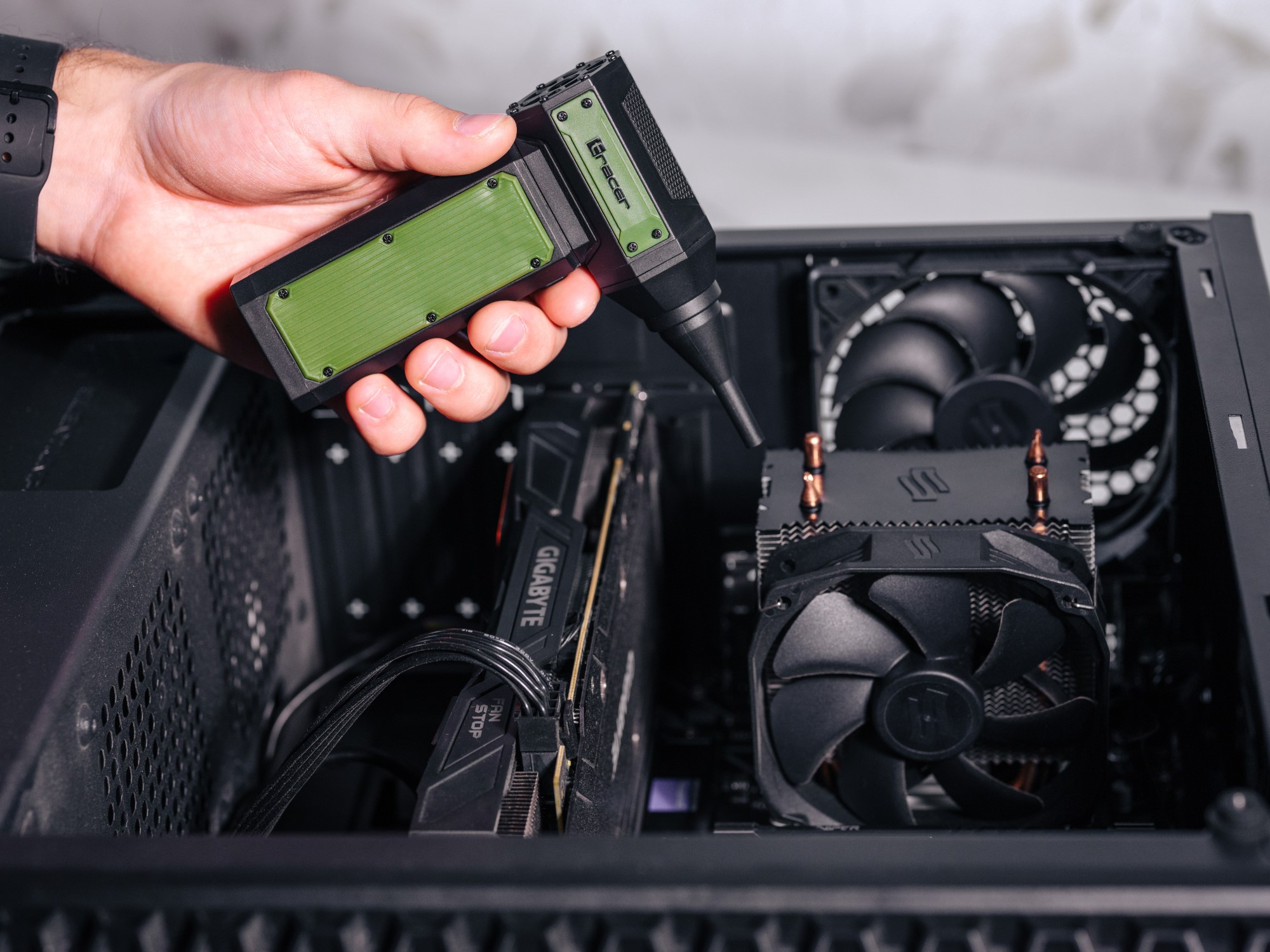 Dłoń trzyma czarno-zieloną dmuchawę powietrza Gracer nad otwartą obudową komputera. W środku znajdują się komponenty, takie jak GPU i chłodnica procesora.