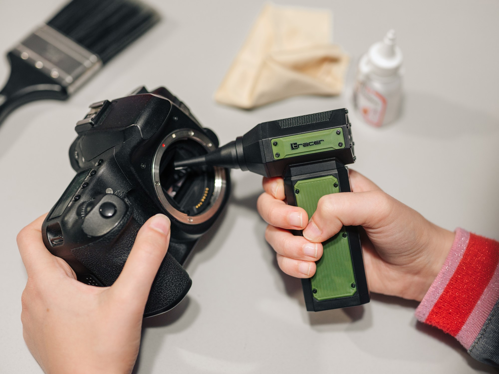 Osoba czyści obiektyw czarnego aparatu za pomocą zielono-czarnego narzędzia czyszczącego. Widoczne są inne przedmioty, takie jak pędzel.